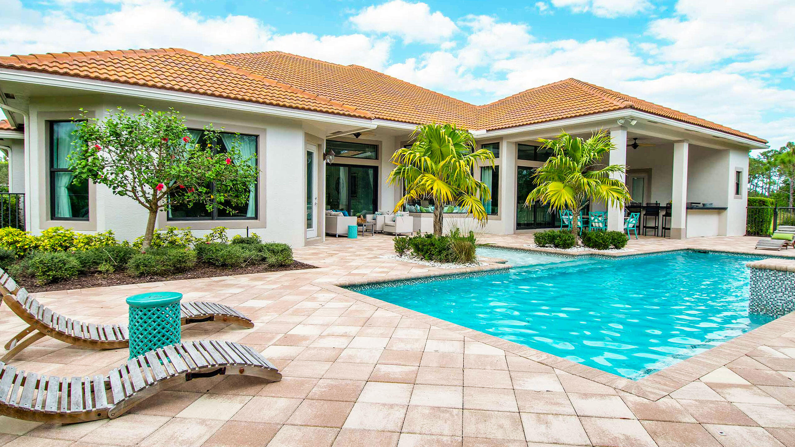 Home’s rear patio and swimming pool with palm trees, lounge chairs, and terracotta roof.