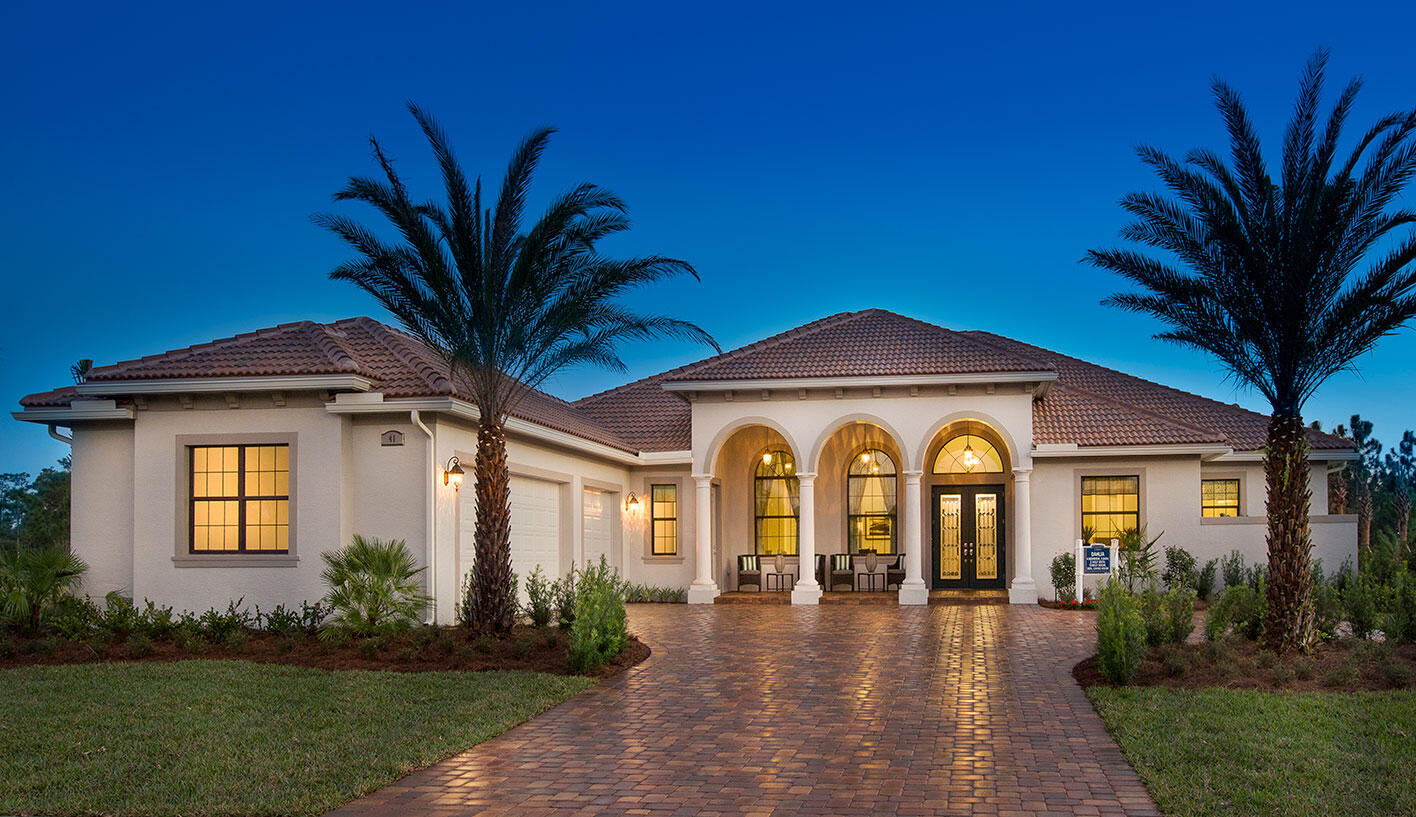 Stucco home with a brick-paver driveway, palm trees, and an illuminated three-arched entrance at dusk.