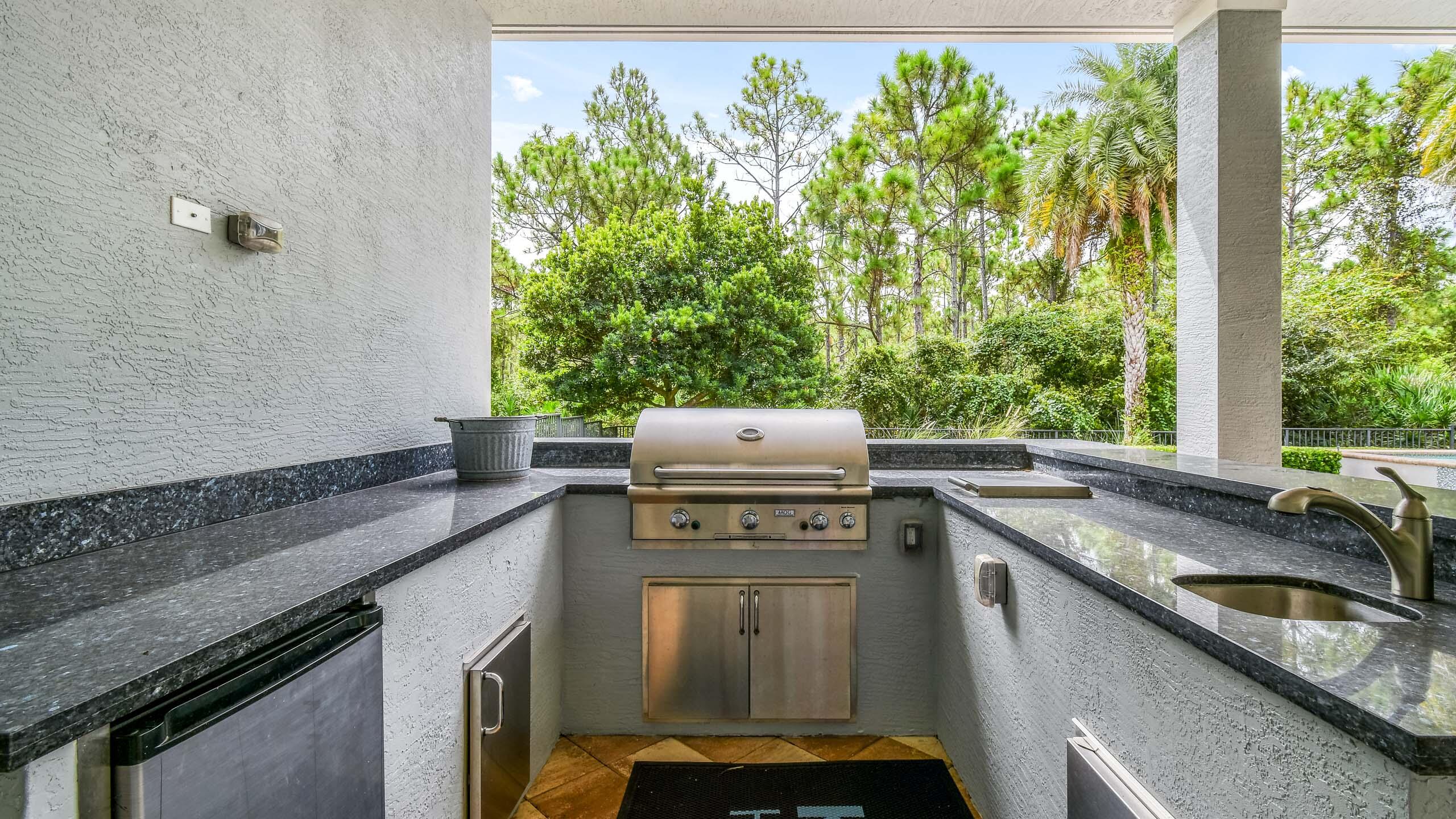 Covered outdoor kitchen with built-in grill, stone counters, sink, and a wooded view.