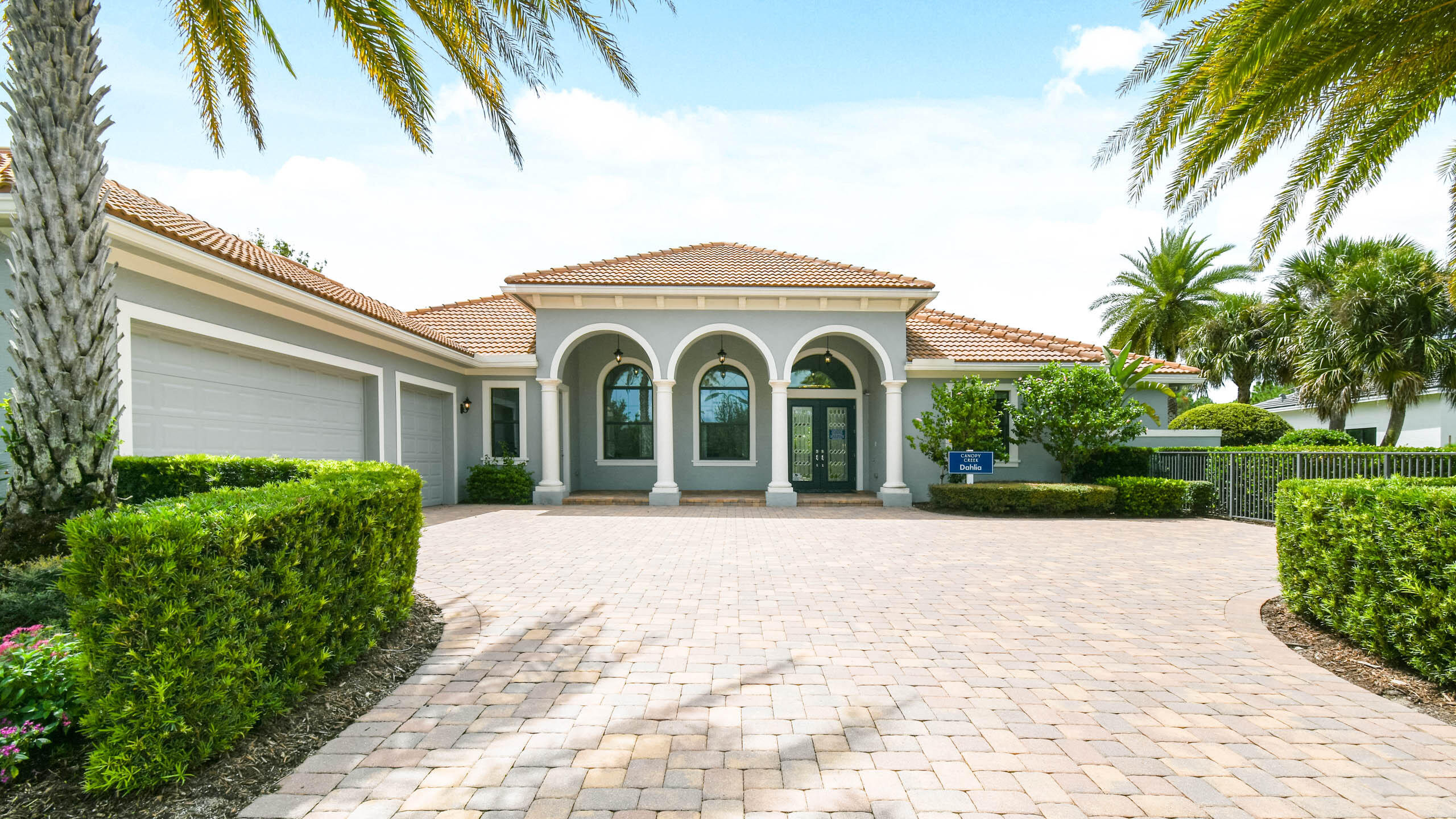 Gray stucco home with arched entryways, terracotta roof, and a broad paver driveway framed by palms.