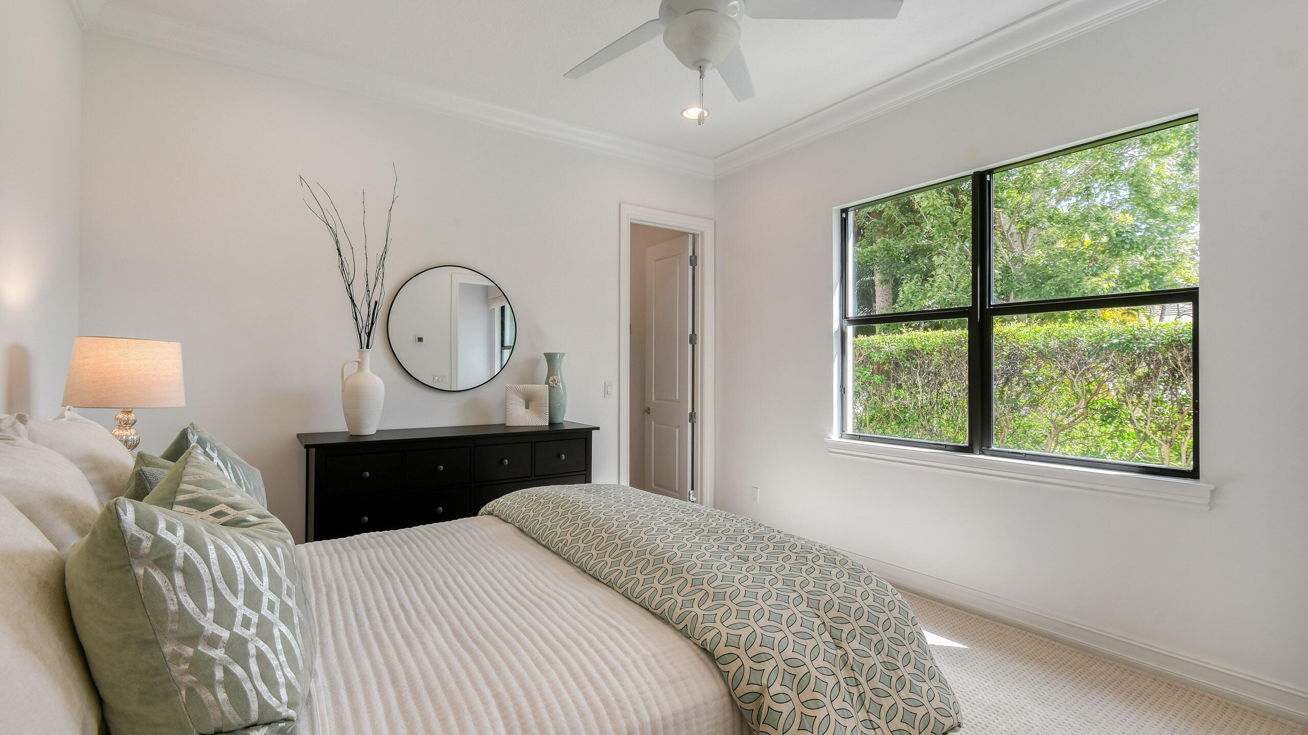 Furnished bedroom with sage-patterned bedding, a black dresser, and a large window overlooking greenery.