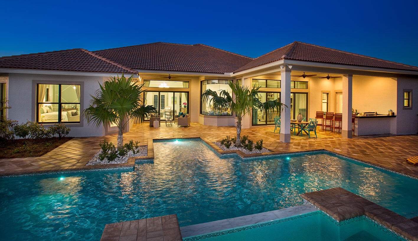Home’s rear patio and covered lanai overlook an illuminated swimming pool at dusk.