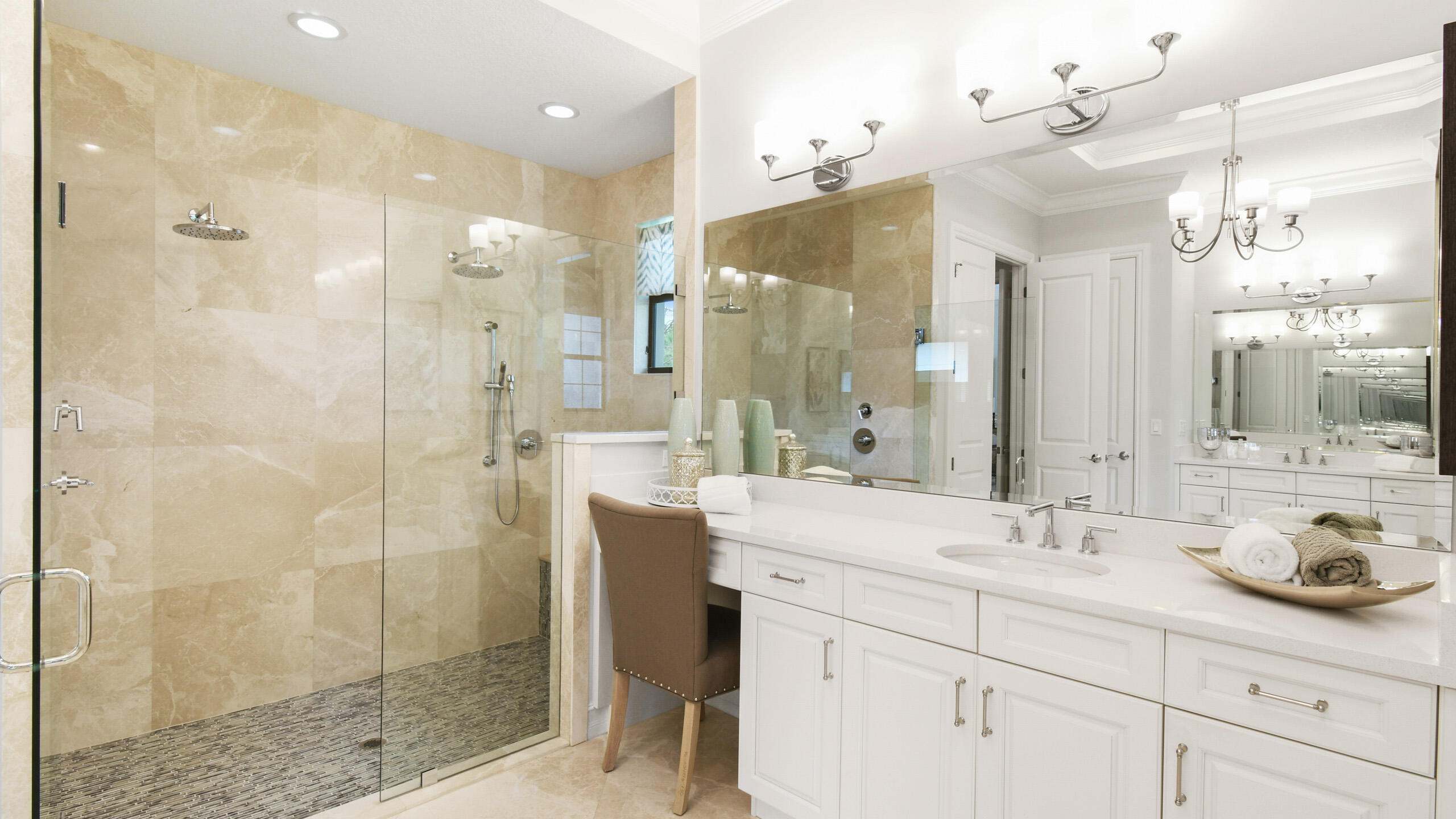 Primary bathroom with a glass walk-in shower, beige stone tile, and long white vanity beneath large mirrors.
