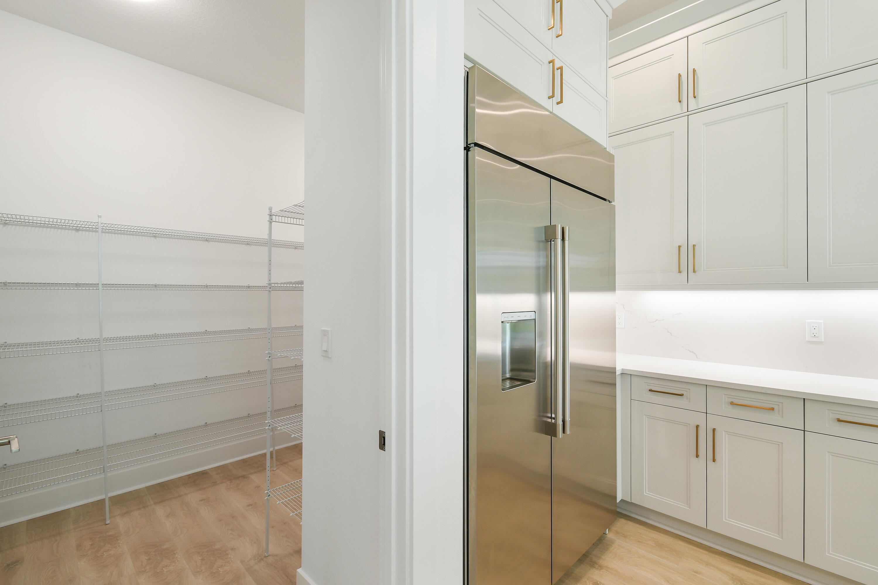 Walk-in pantry with wire shelving beside a kitchen with a stainless steel refrigerator and white cabinetry.