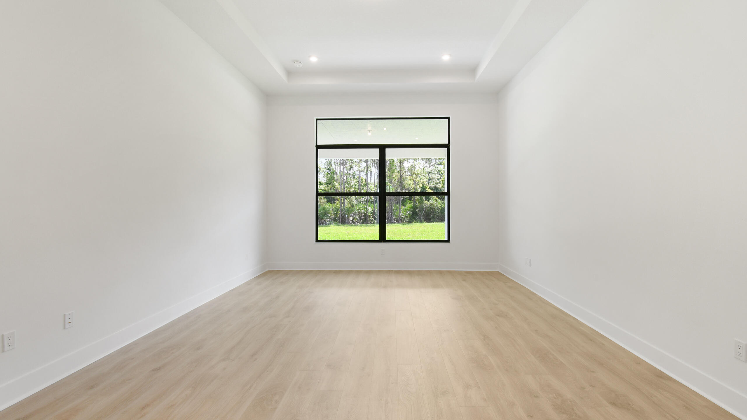 Empty room with pale wood floors and a large black-framed window overlooking a green lawn and trees.