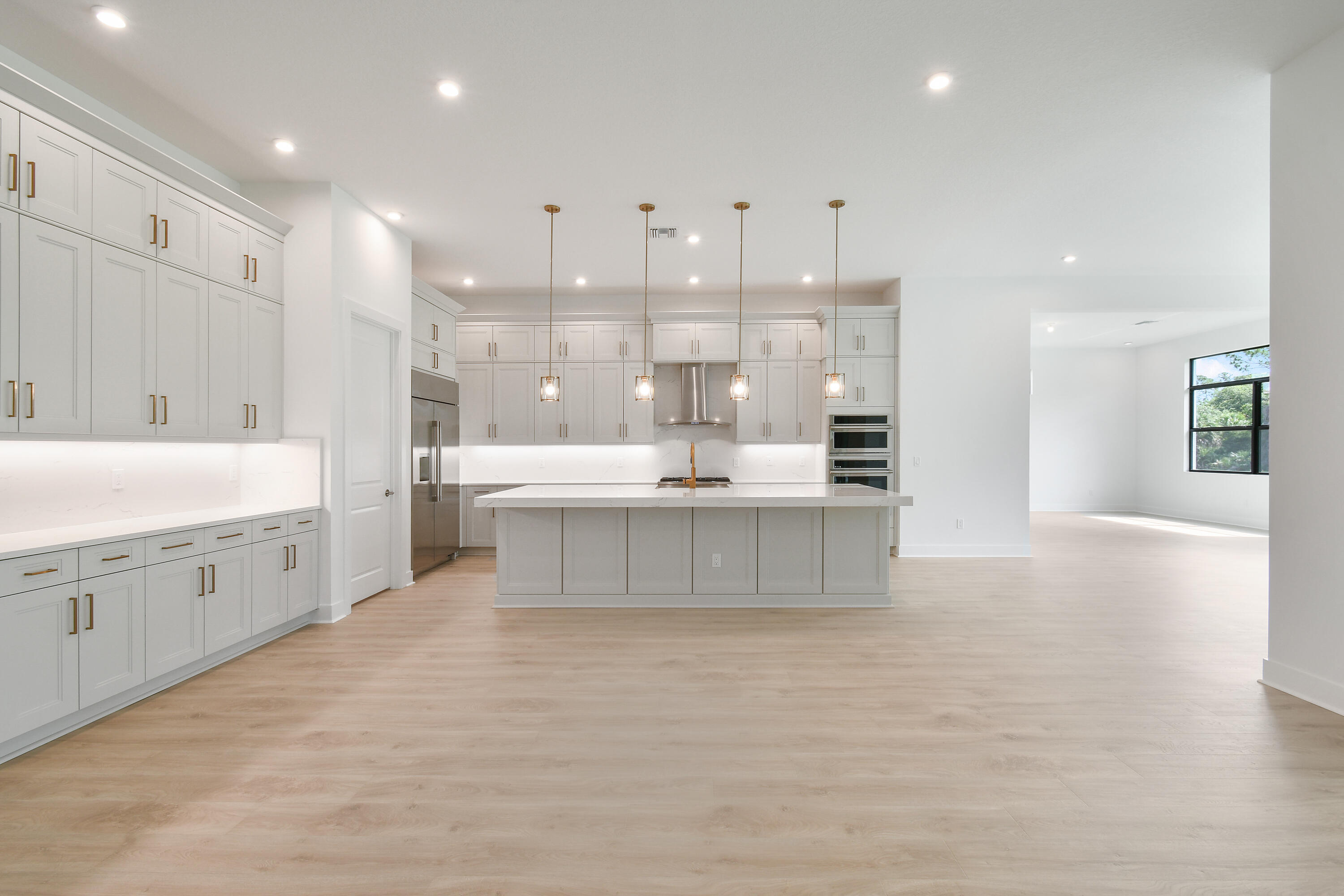 Vacant white kitchen with a large island, pendant lights, stainless appliances, and light wood floors.