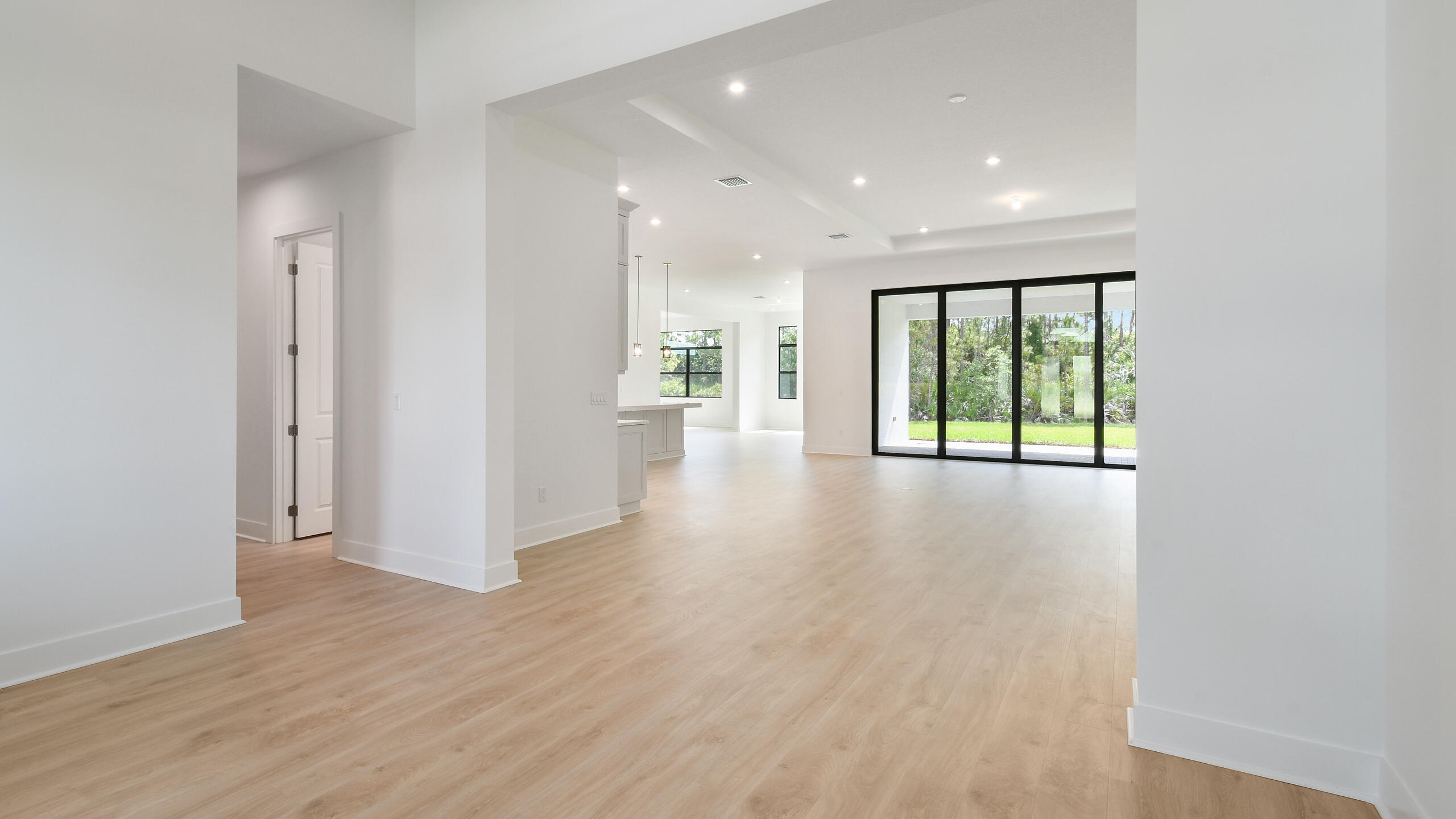 Empty open-plan living area with light wood floors and black-framed sliding doors to the yard.