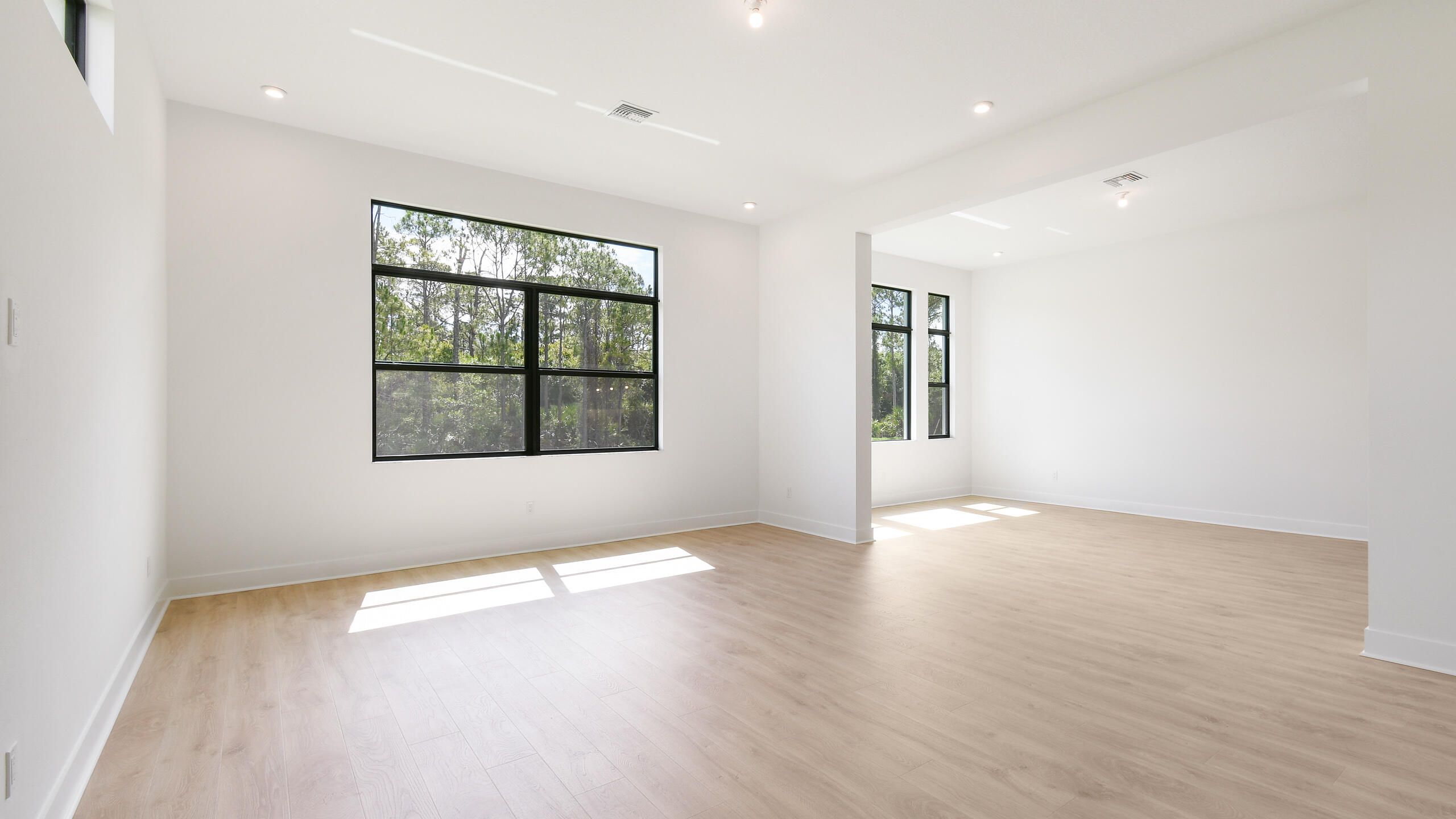 Vacant open living area with pale wood floors, high ceilings, and large black-framed windows.