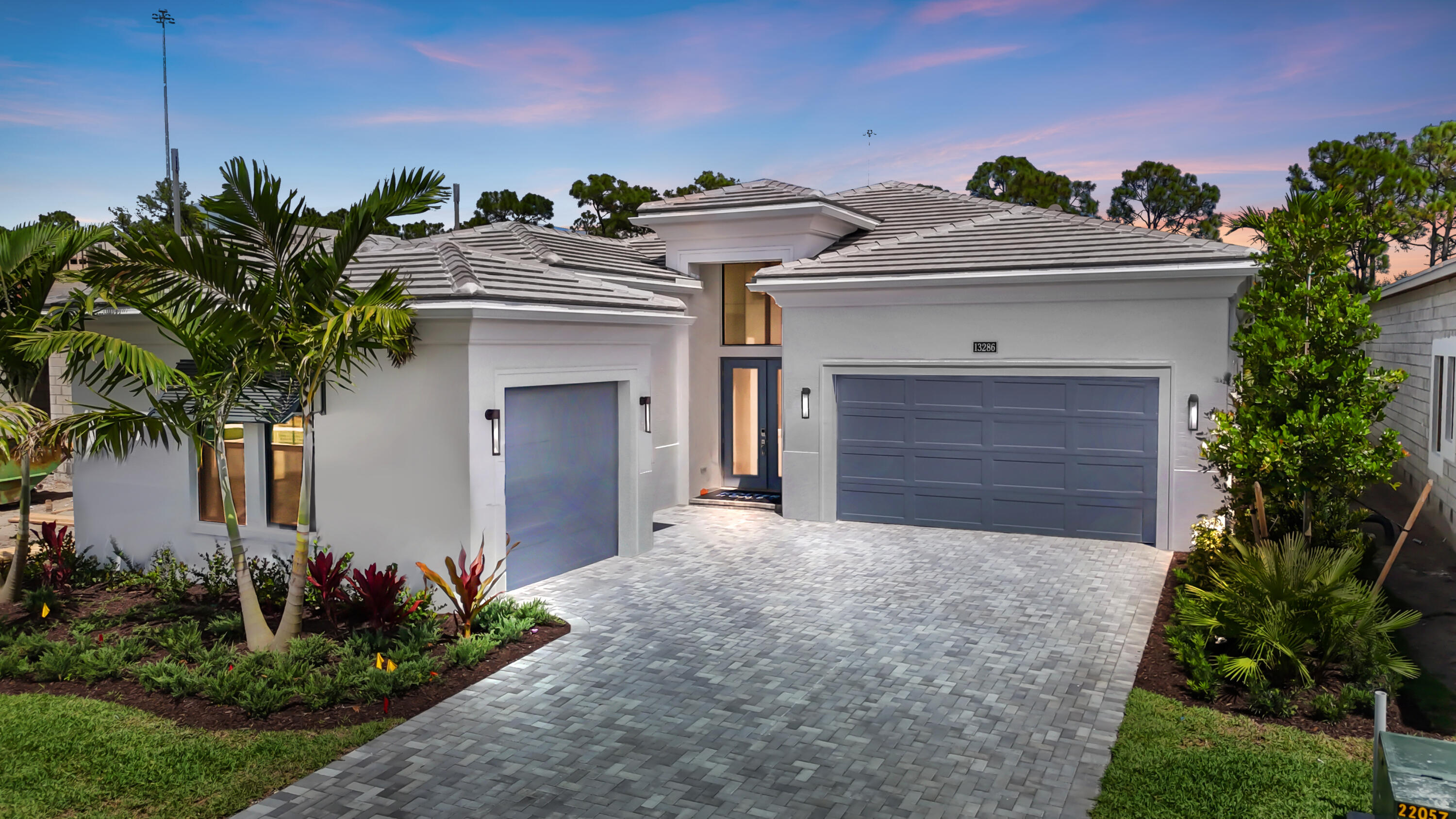Modern white home with two blue-gray garage doors, tropical landscaping, and a wide paver driveway at dusk.