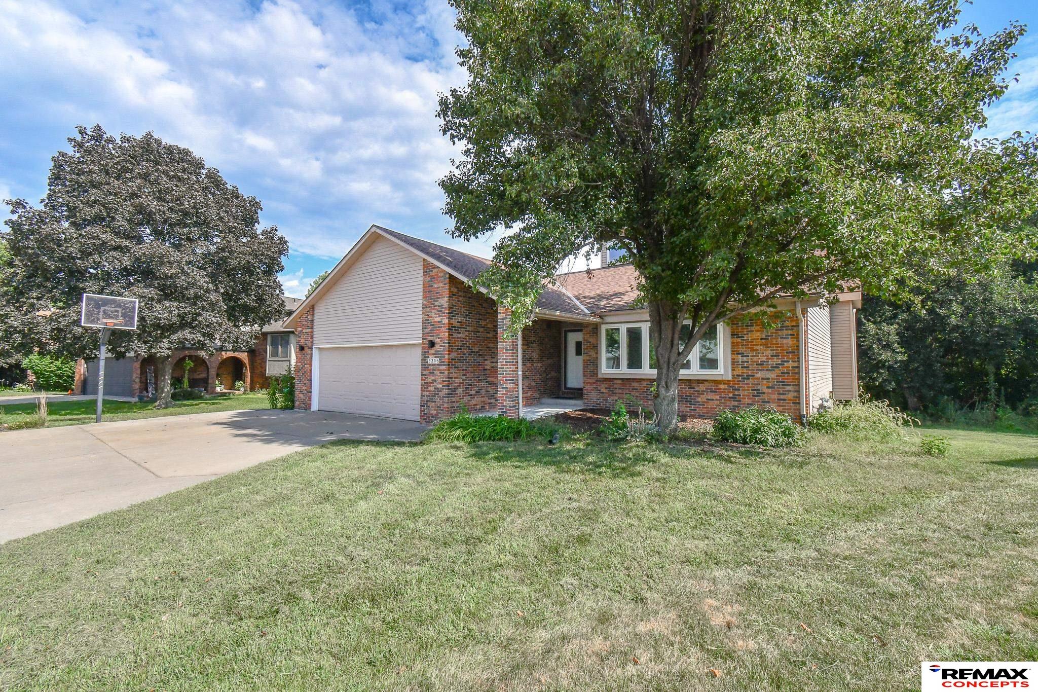 Brick home with a two-car garage, shaded by a large tree beside the driveway and basketball hoop.