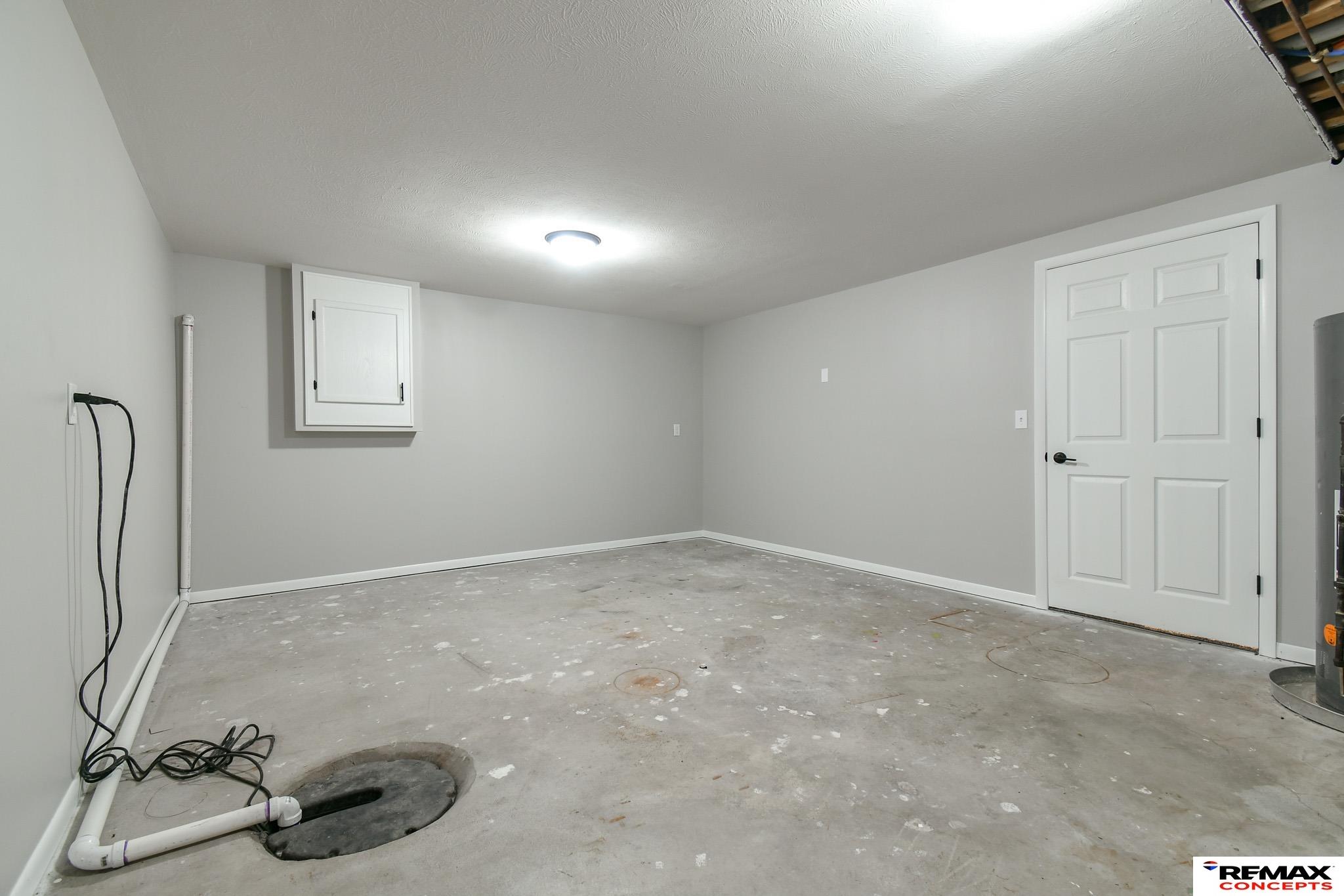 Unfinished basement utility room with a concrete floor, sump pit, exposed plumbing, and white paneled door.