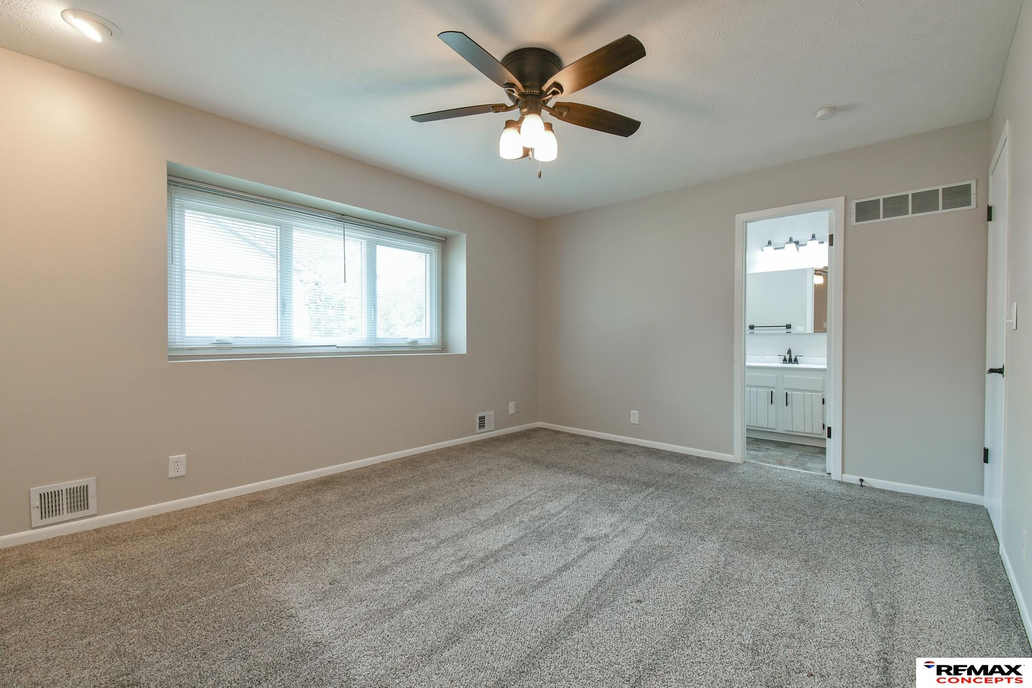 Empty carpeted bedroom with a wide window, ceiling fan, and doorway to a bathroom.