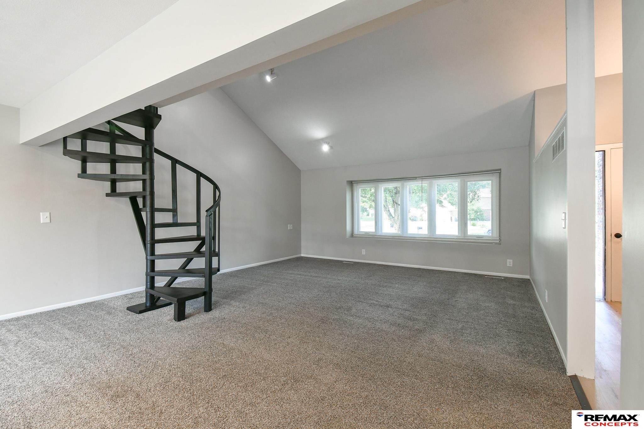 Empty living room with a black spiral staircase, gray carpet, and a wide window.
