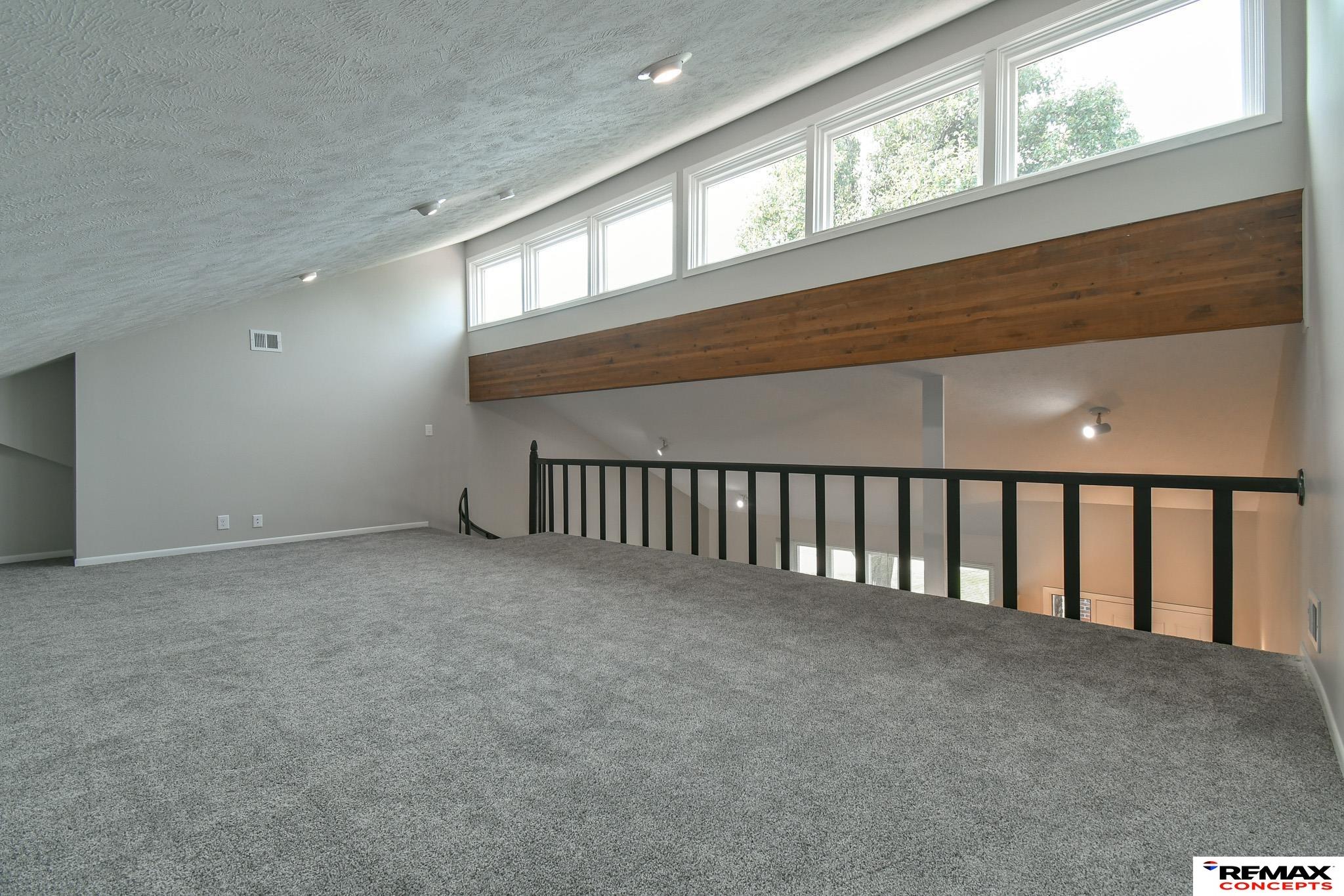 Carpeted loft with a black railing, sloped ceiling, clerestory windows, and a broad wood-paneled beam.