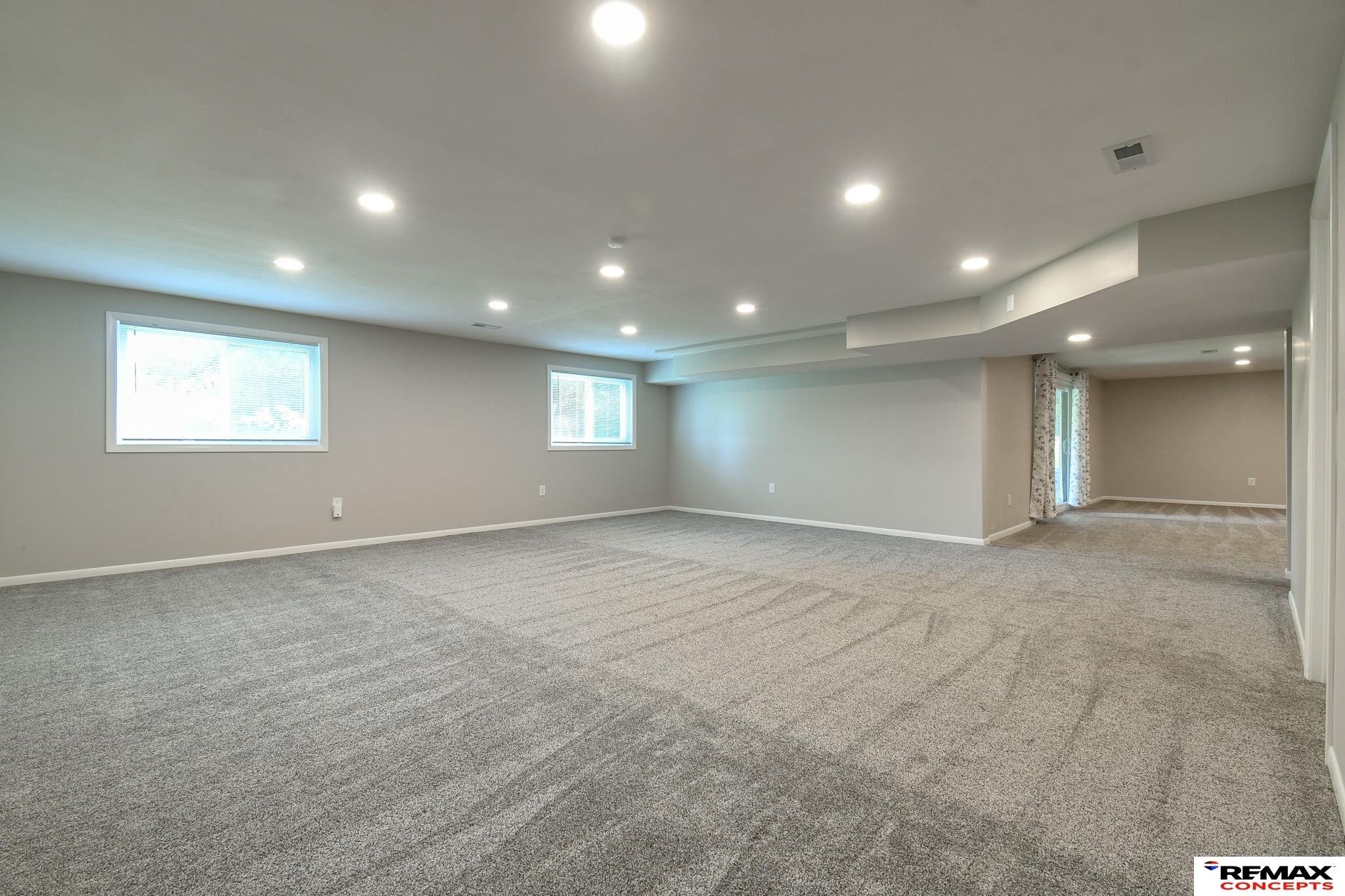 Vacant carpeted basement recreation room with recessed lights and an adjoining room.