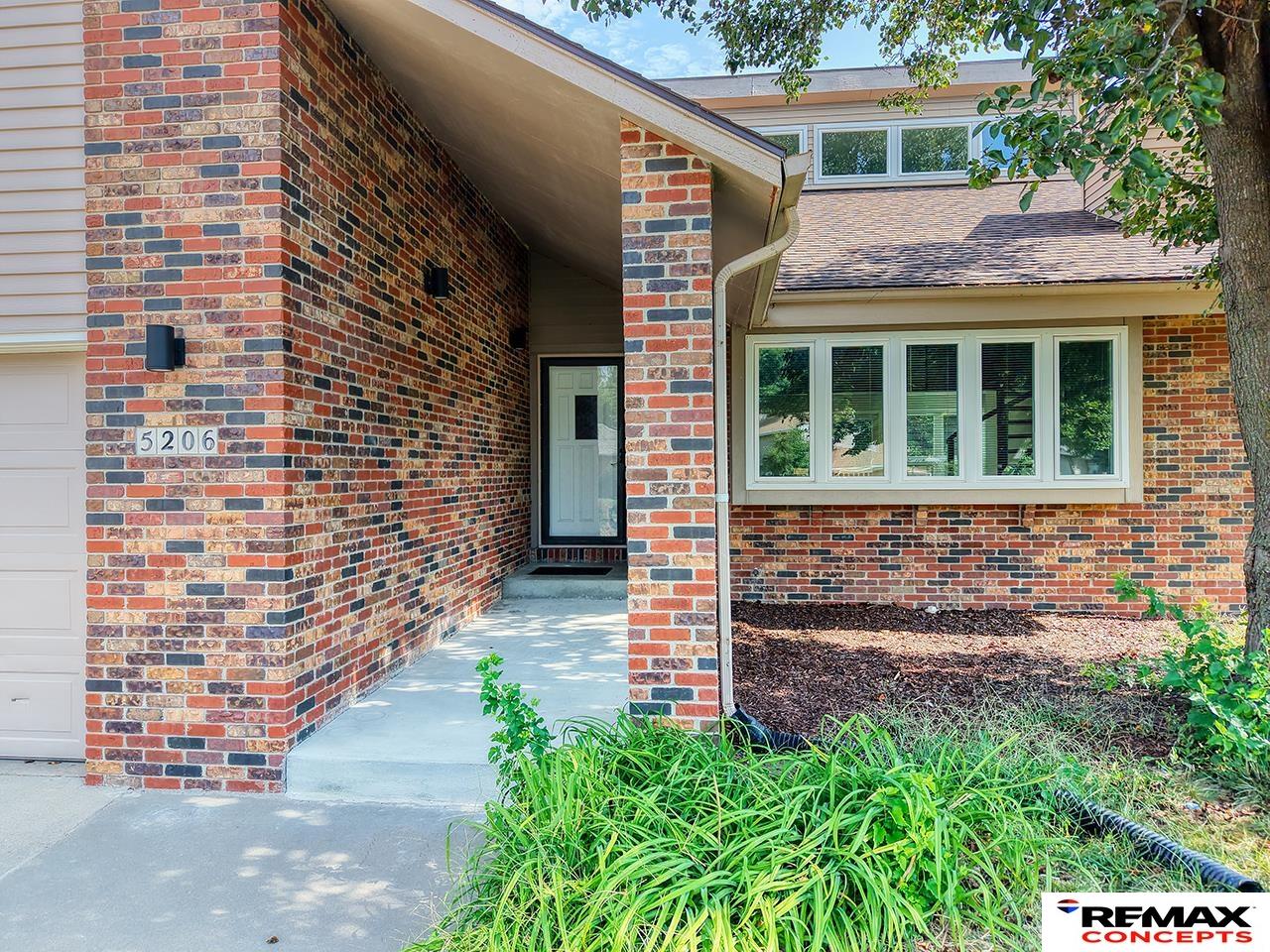 Brick home entrance with a covered walkway, white-framed windows, and leafy landscaping.
