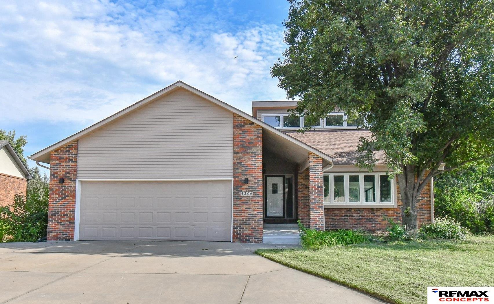 Home exterior with a two-car garage, red brick accents, and a large tree beside the driveway.