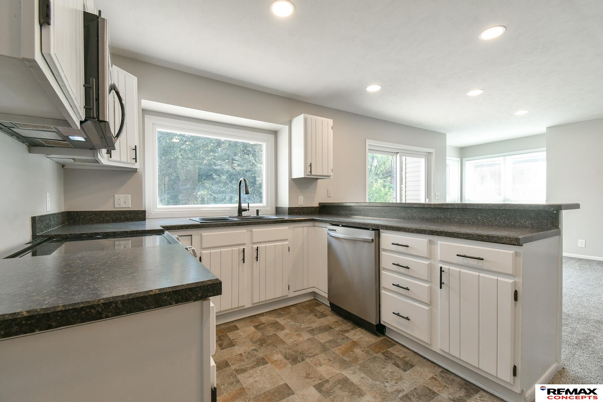 Kitchen with white cabinets, dark countertops, stainless steel appliances, and a large window over the sink.
