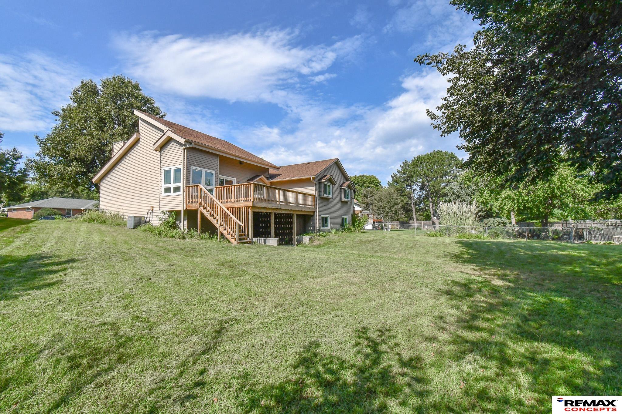 Rear of a beige-sided house with a raised wooden deck, stairs, and a broad tree-lined lawn.