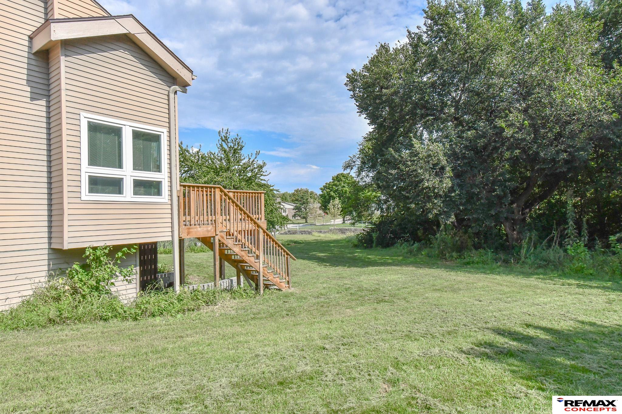 Home exterior with wooden deck stairs beside a broad lawn and mature tree.