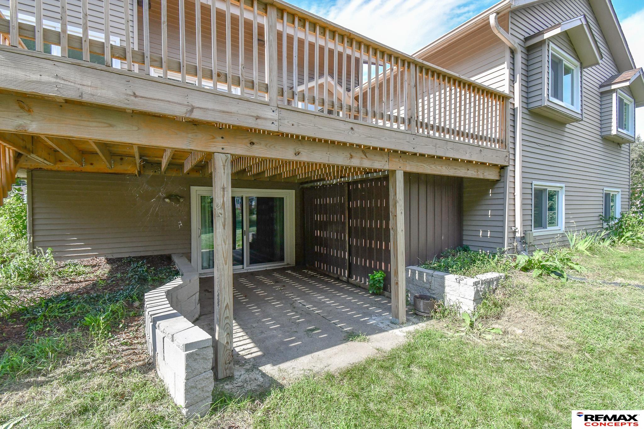 Shaded concrete patio beneath a large elevated wooden deck at a gray-sided house.