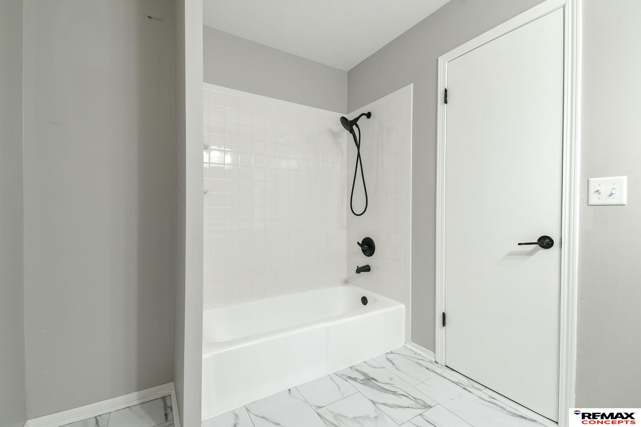 Bathroom with a white tiled tub and shower, black fixtures, and marble-look flooring.
