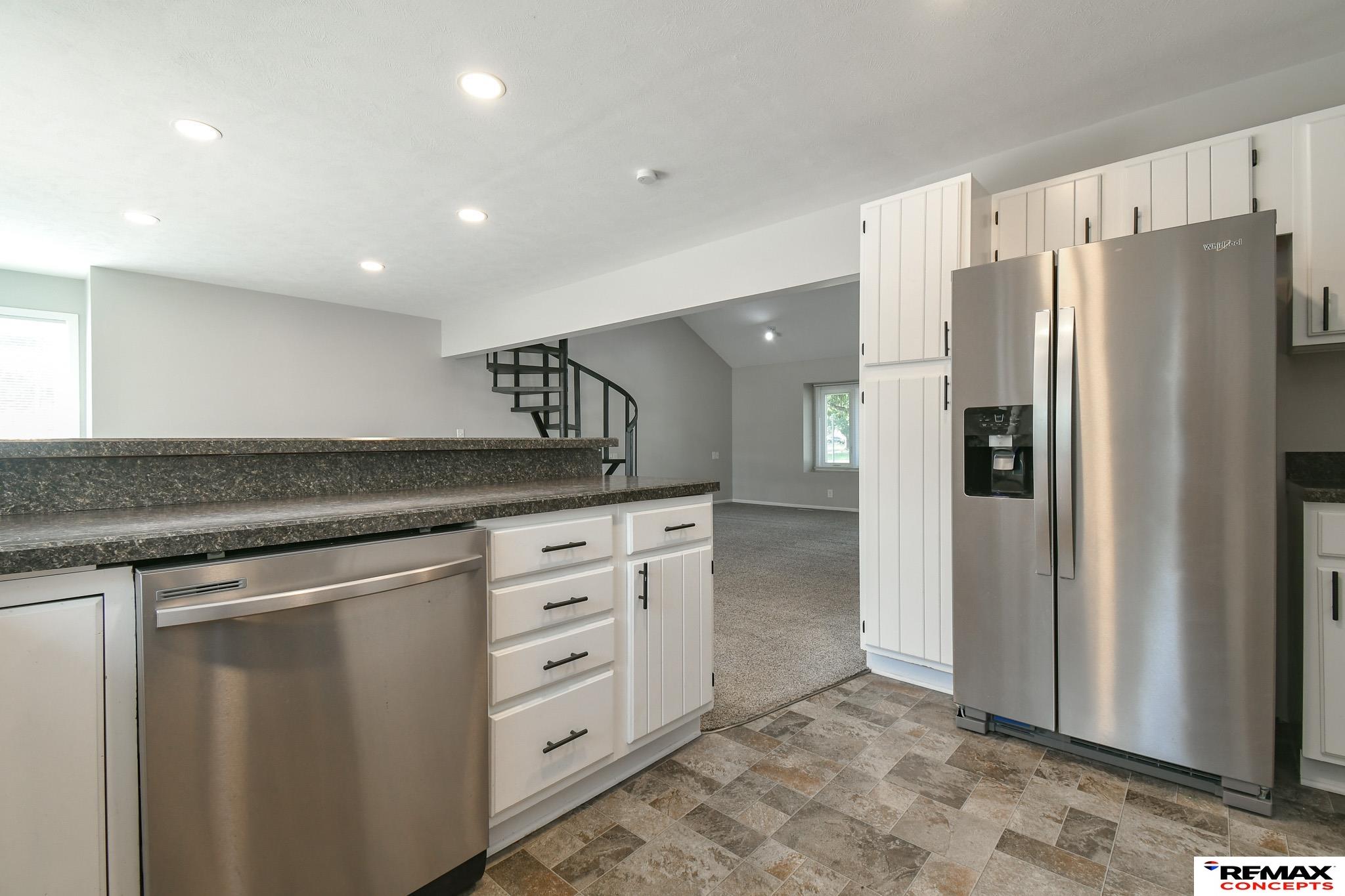 Kitchen with white cabinets, dark stone counters, stainless steel appliances, and a curved staircase beyond.