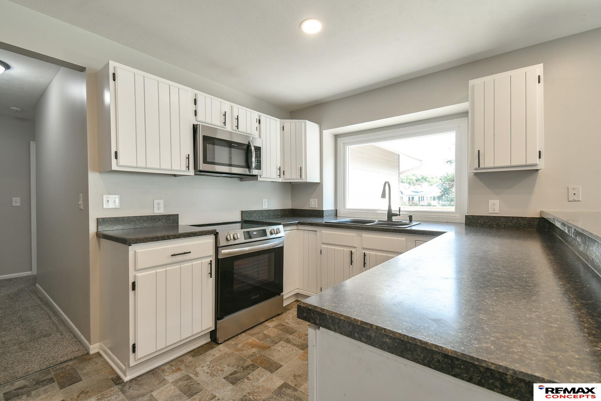 Kitchen with white paneled cabinets, dark countertops, stainless steel appliances, and a large window over the sink.