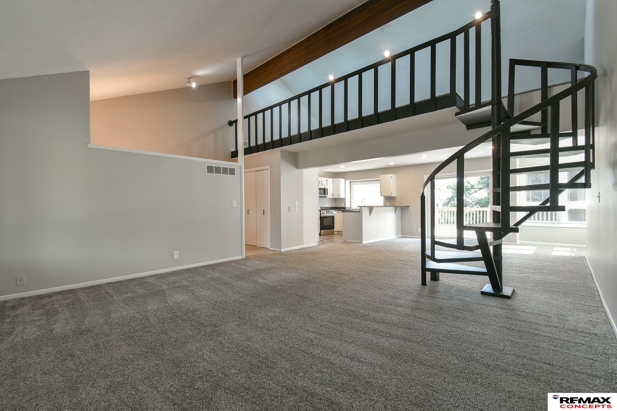 Vacant living room with a black spiral staircase, open mezzanine, and kitchen beyond.