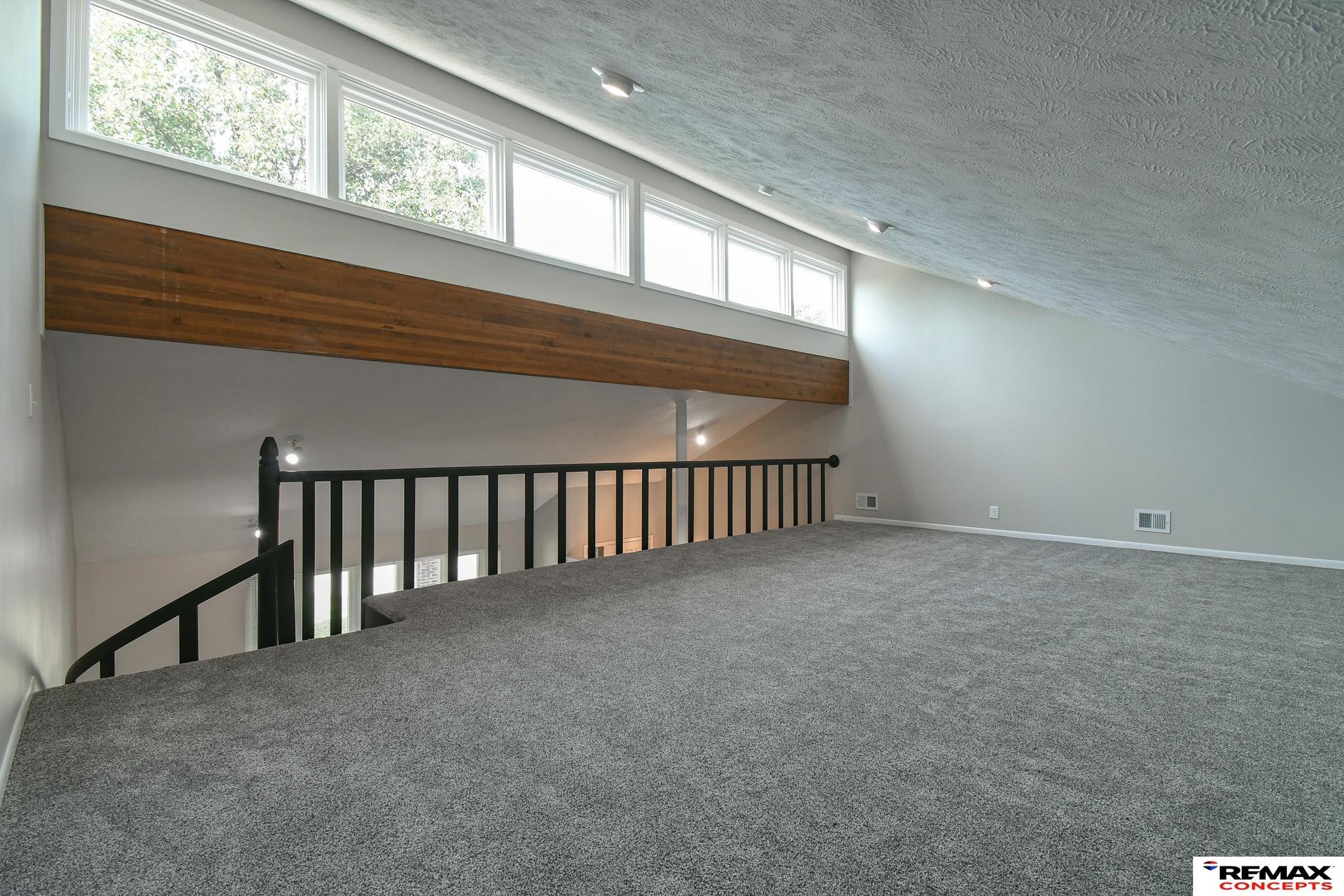 Carpeted loft with black railing, high sloped ceiling, and a row of upper windows.