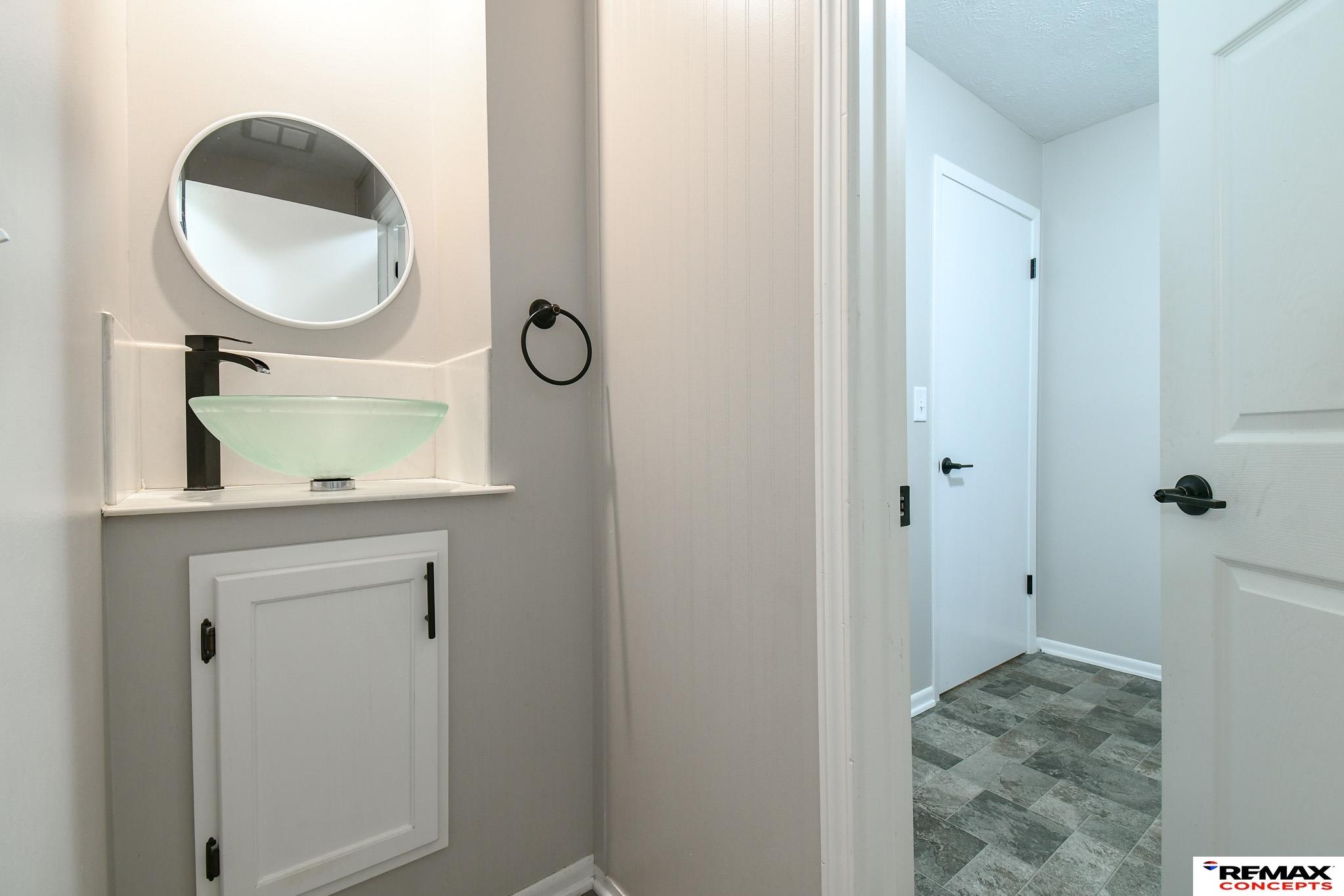 Bathroom vanity with a pale green glass vessel sink and round mirror beside a doorway to gray-tiled flooring.