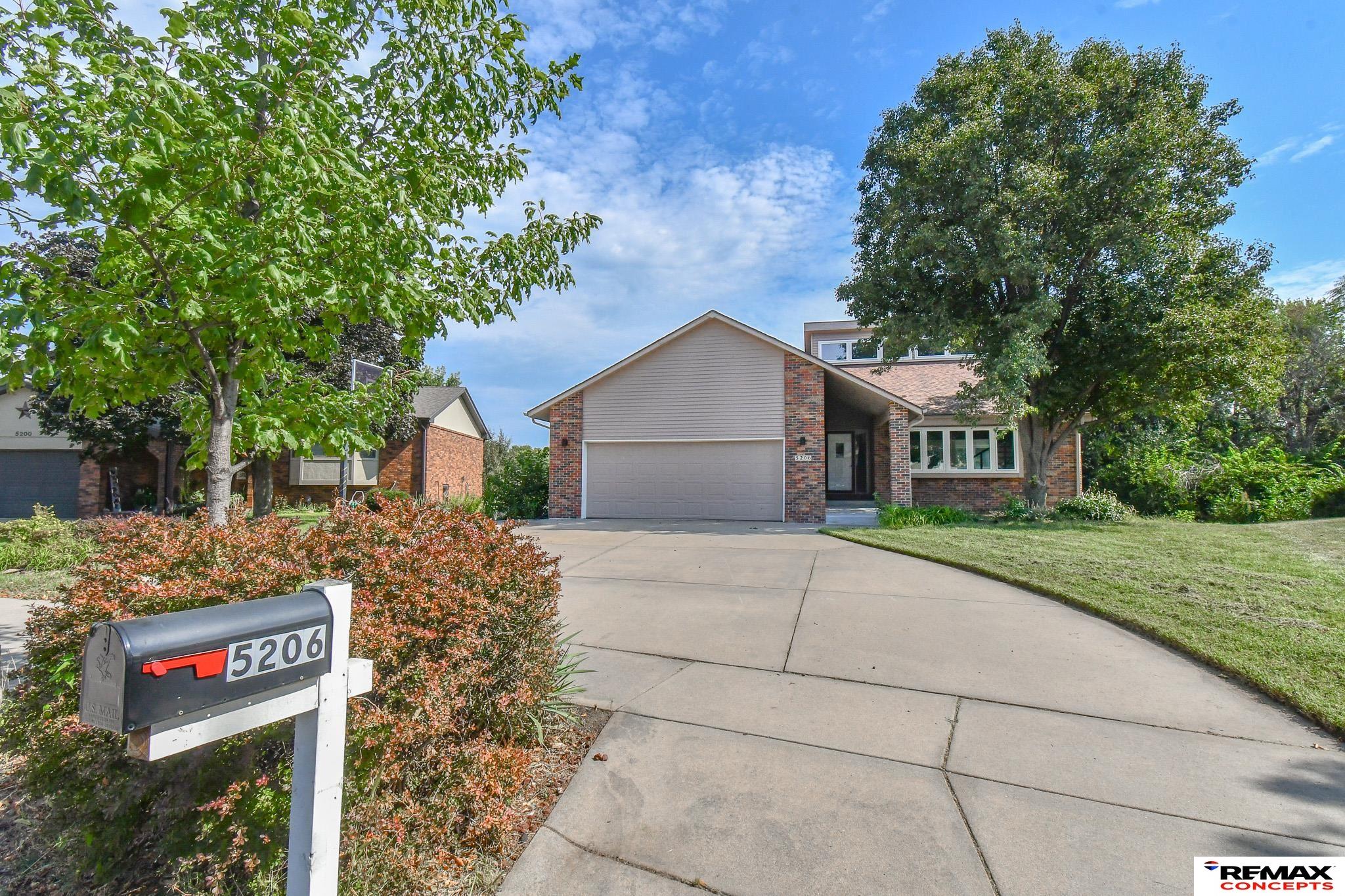 Brick-and-siding house with a double garage, broad driveway, mature trees, and a 5206 mailbox.