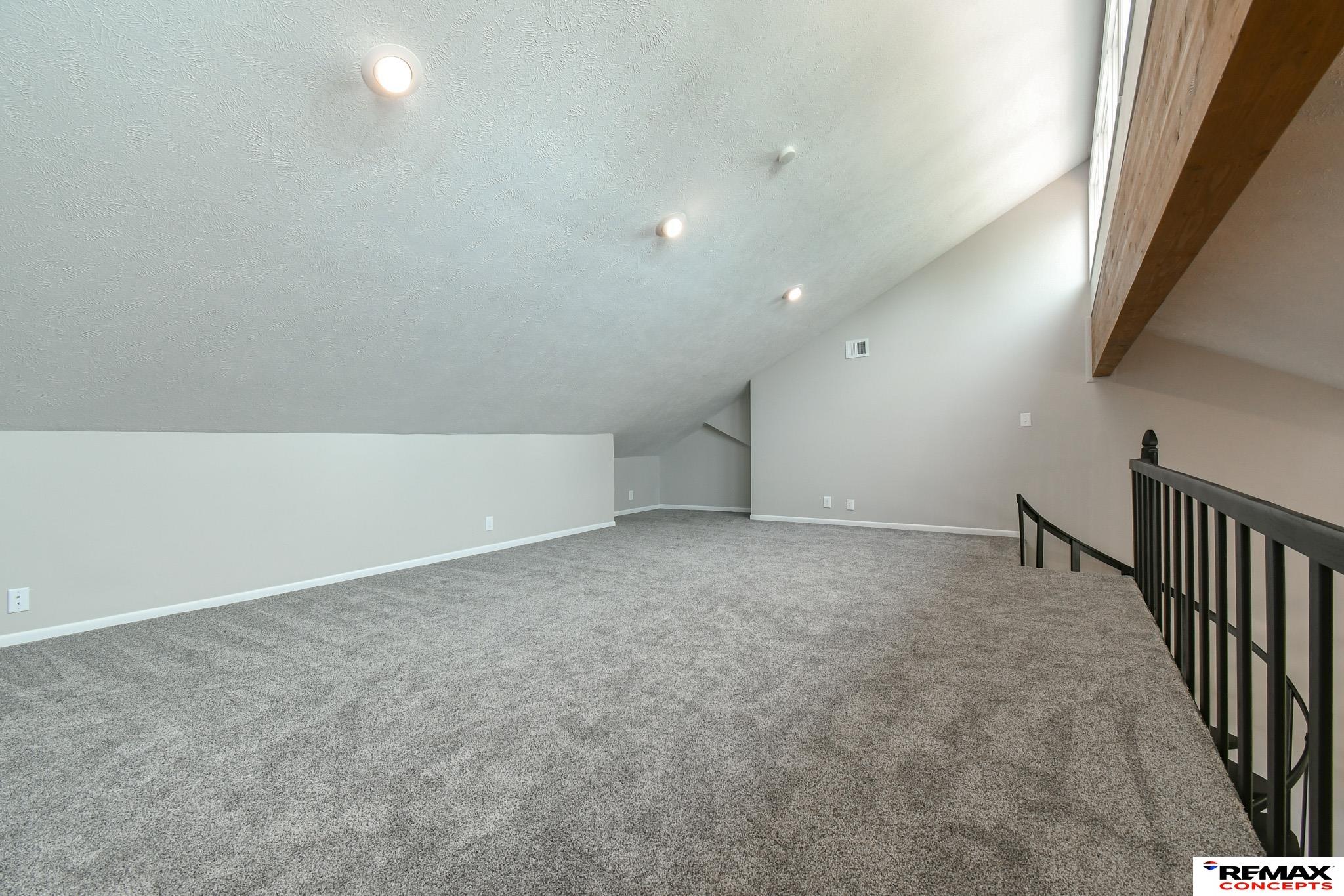 Carpeted loft with a sloped ceiling, exposed wood beam, and black railing.