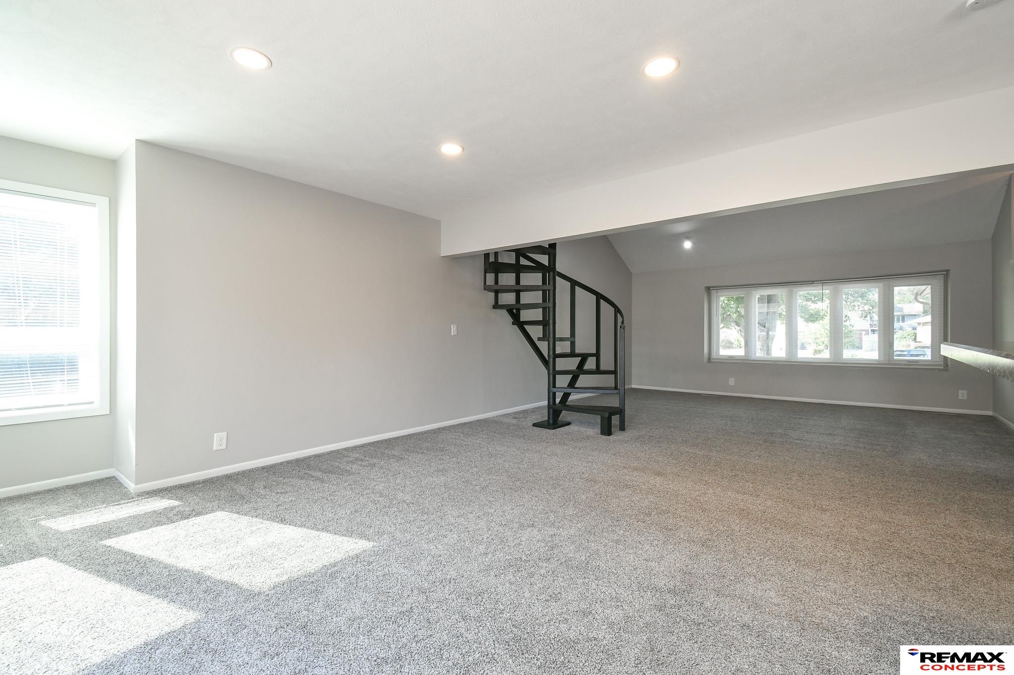 Vacant carpeted living area with a black spiral staircase and wide windows.