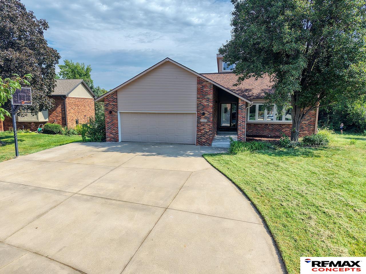 Brick house with an attached two-car garage, broad driveway, mature trees, and a basketball hoop.