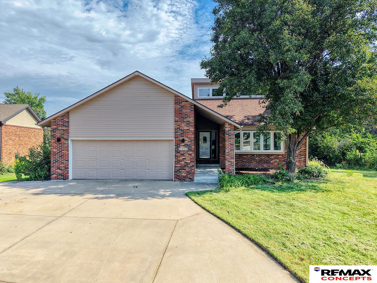 Brick-and-siding house with a two-car garage, wide driveway, and mature tree in the front lawn.
