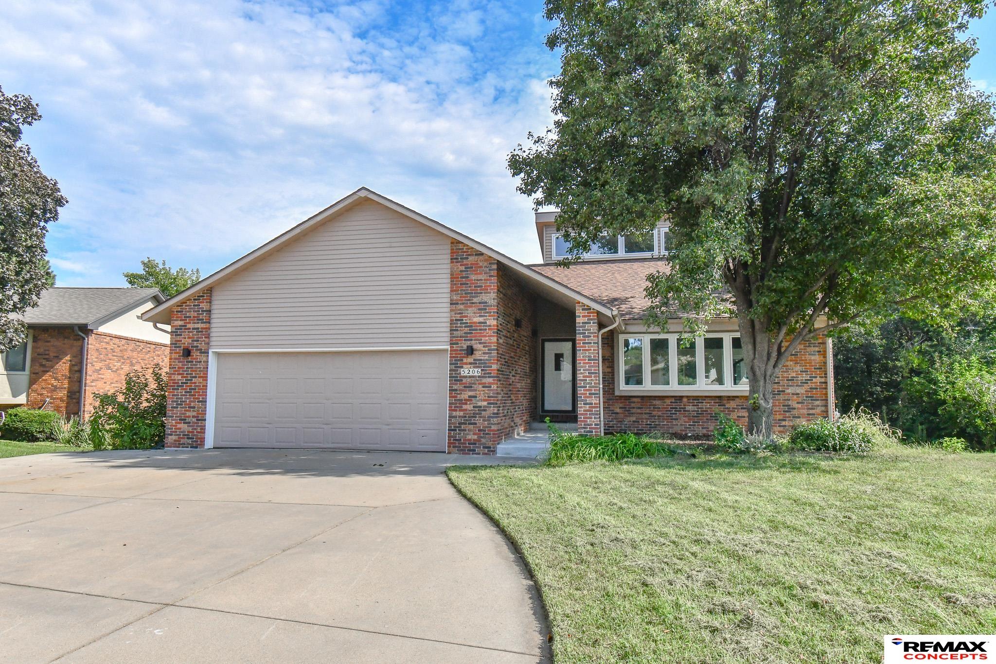 Brick-and-siding home with a two-car garage, broad driveway, and mature tree shading the front lawn.