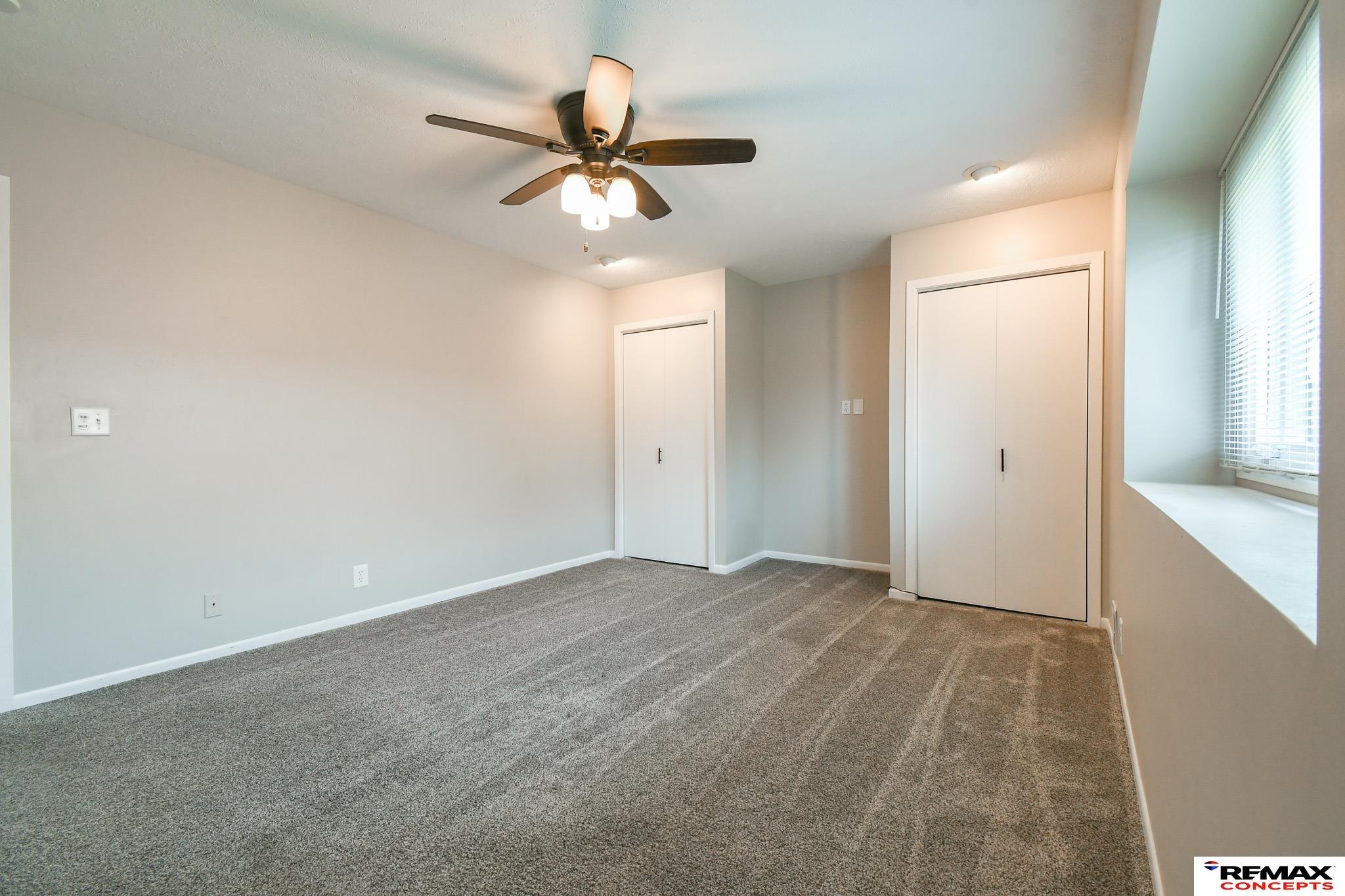 Empty bedroom with gray carpet, two closets, a ceiling fan, and a large window with blinds.