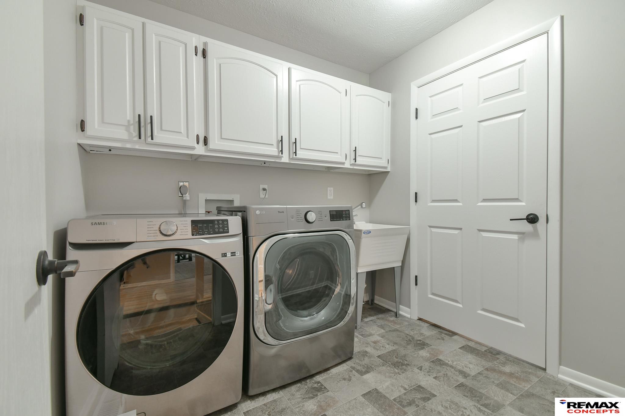 Laundry room with front-loading washer and dryer, white cabinets, utility sink, and patterned tile floor.