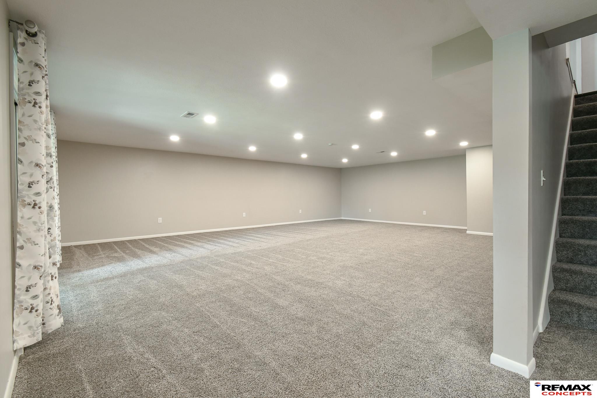 Empty carpeted basement room with recessed lights and a staircase along the right side.
