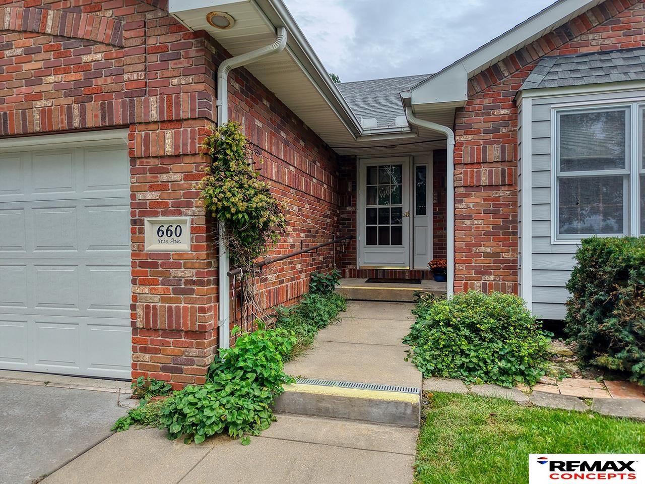 Brick home entrance with a covered walkway, white door, attached garage, and leafy landscaping.