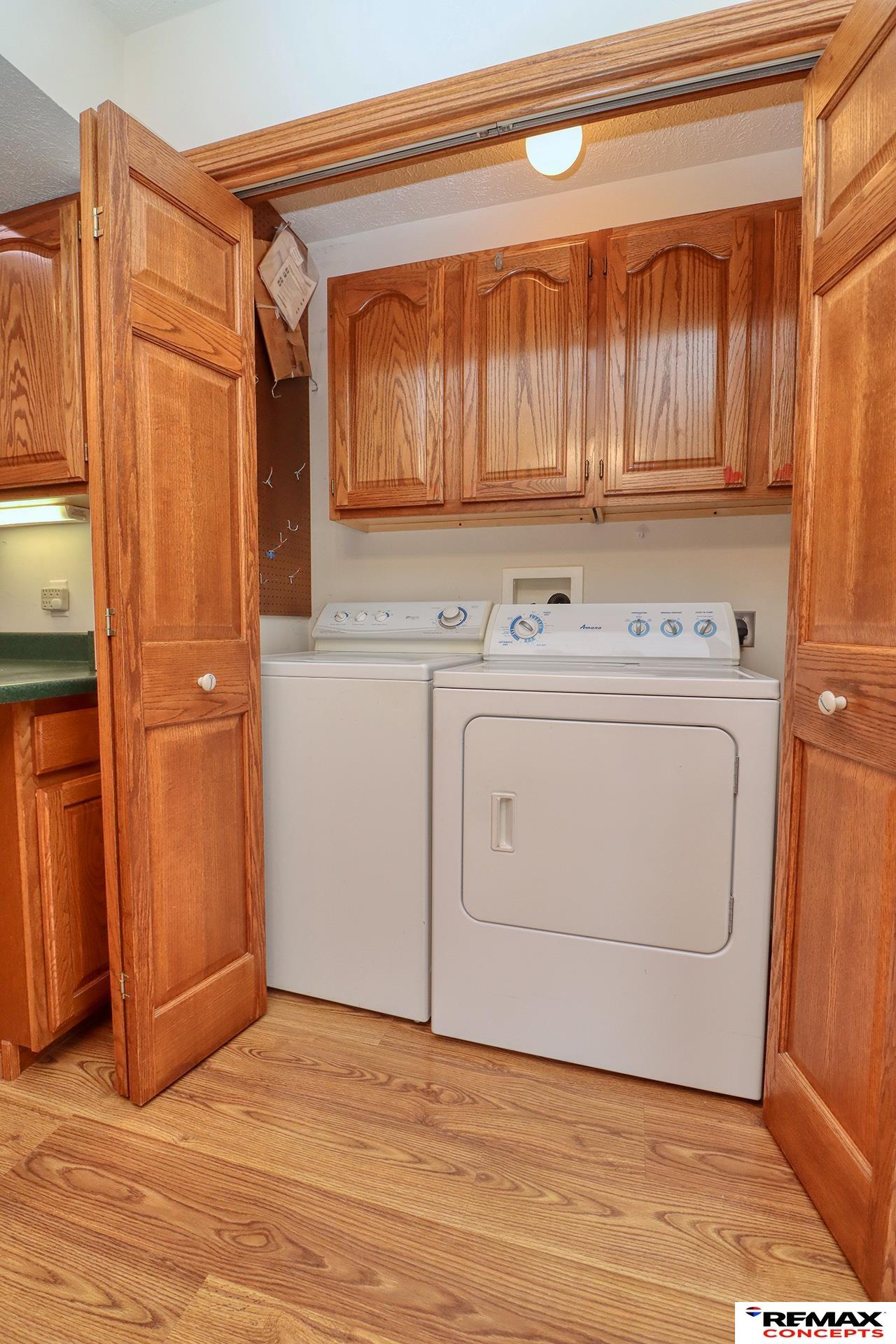 Laundry closet with washer and dryer beneath oak cabinets and open wooden doors.