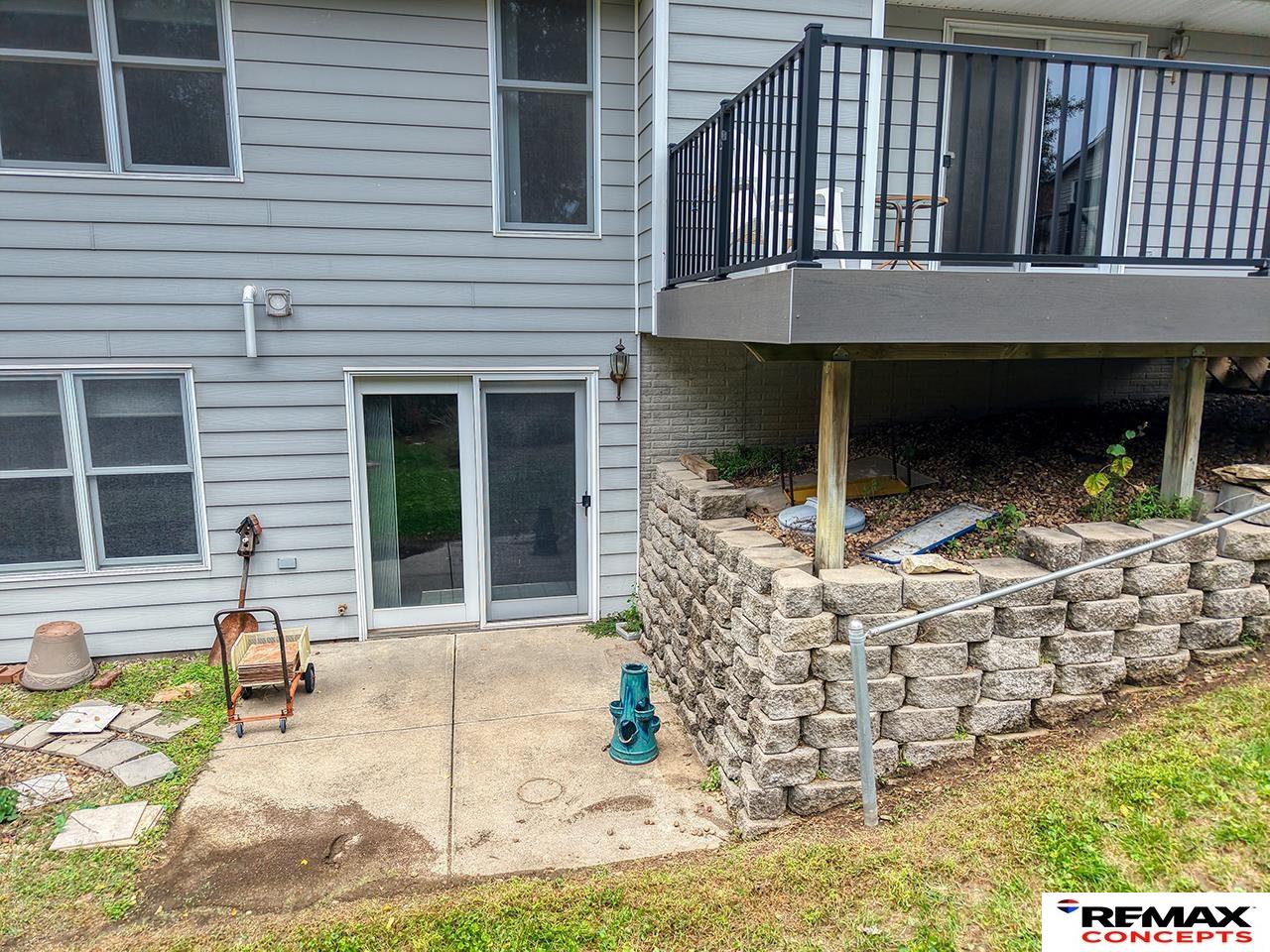 House’s rear deck above a concrete patio, with black railings and a stone retaining wall.