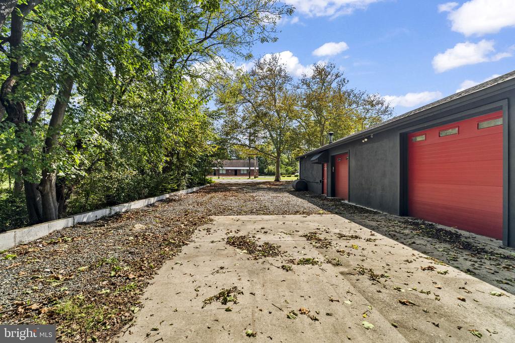 Gray garage with red doors beside a leaf-strewn concrete driveway under mature trees.