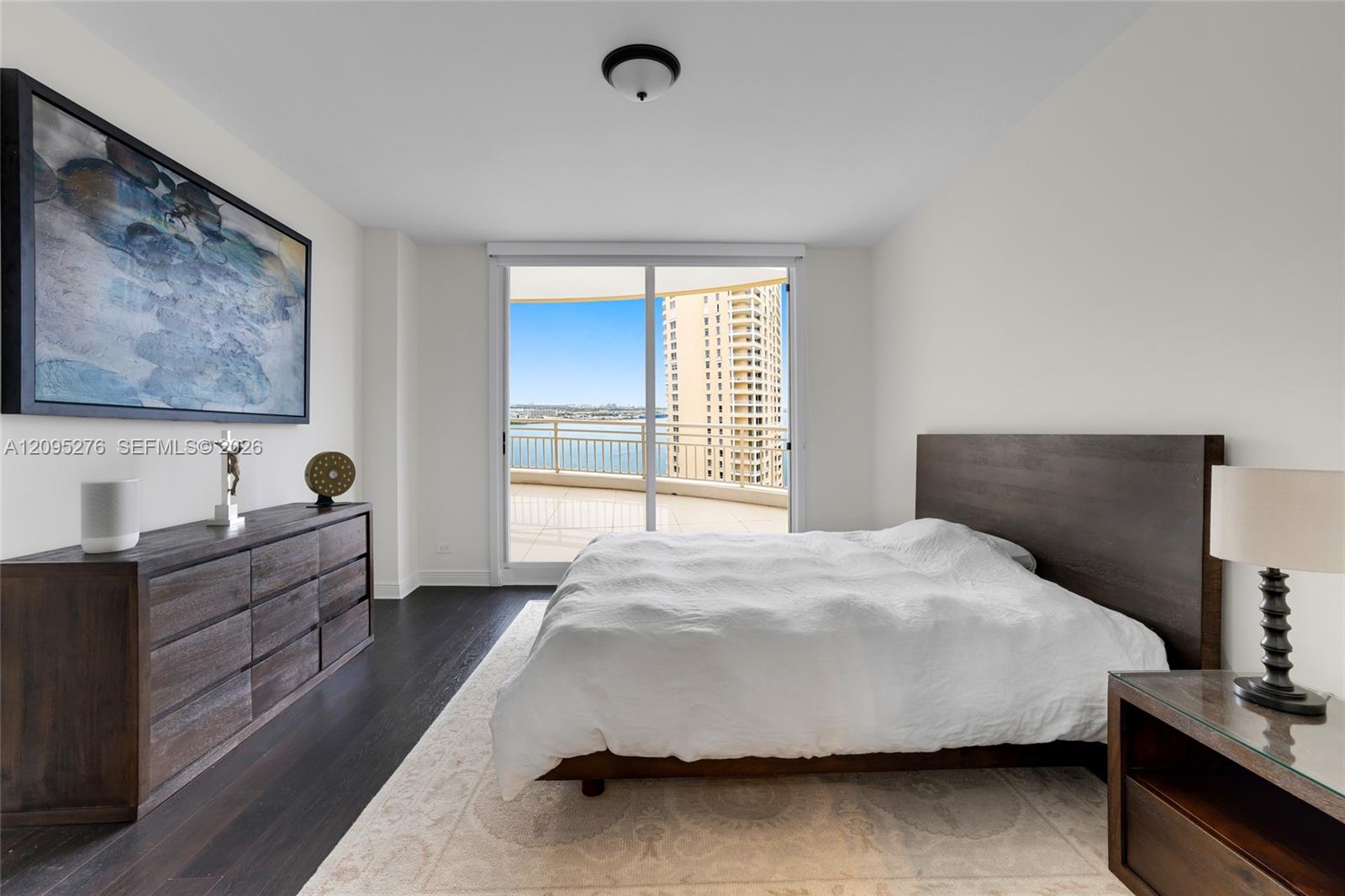 Bedroom with white bedding and a balcony overlooking water and a neighboring high-rise.