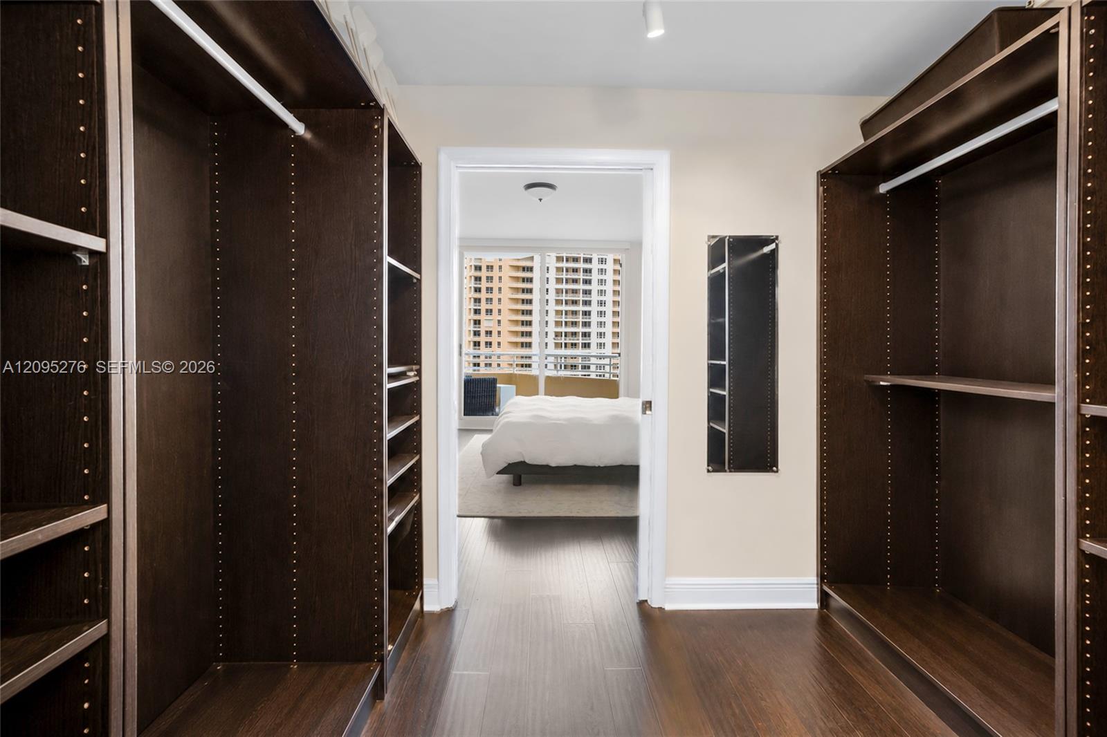 Walk-in closet with dark built-in shelving opening into a bedroom with balcony windows.