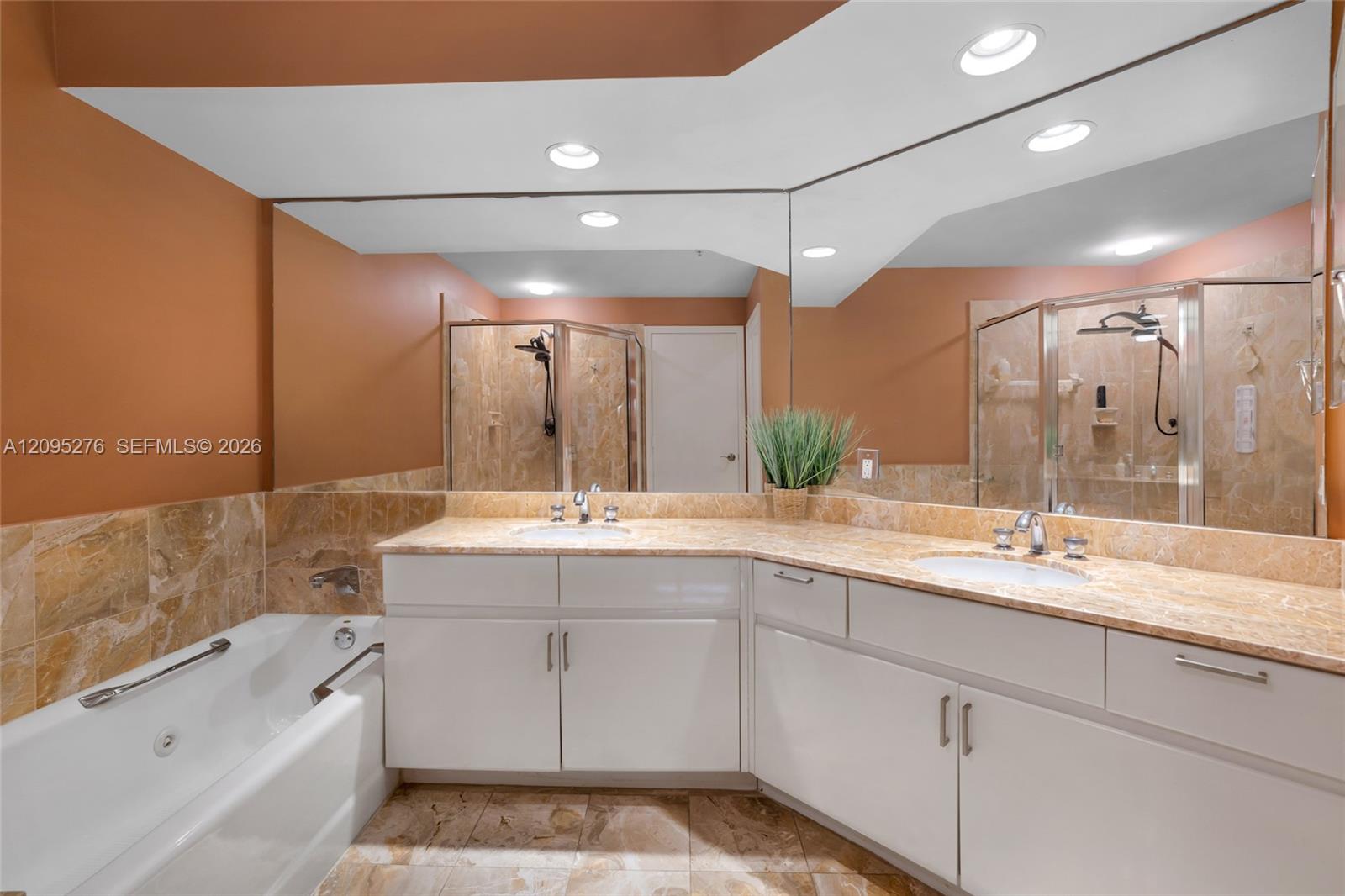 Bathroom with a soaking tub, long dual-sink vanity, stone tile, and mirrored walls reflecting glass showers.
