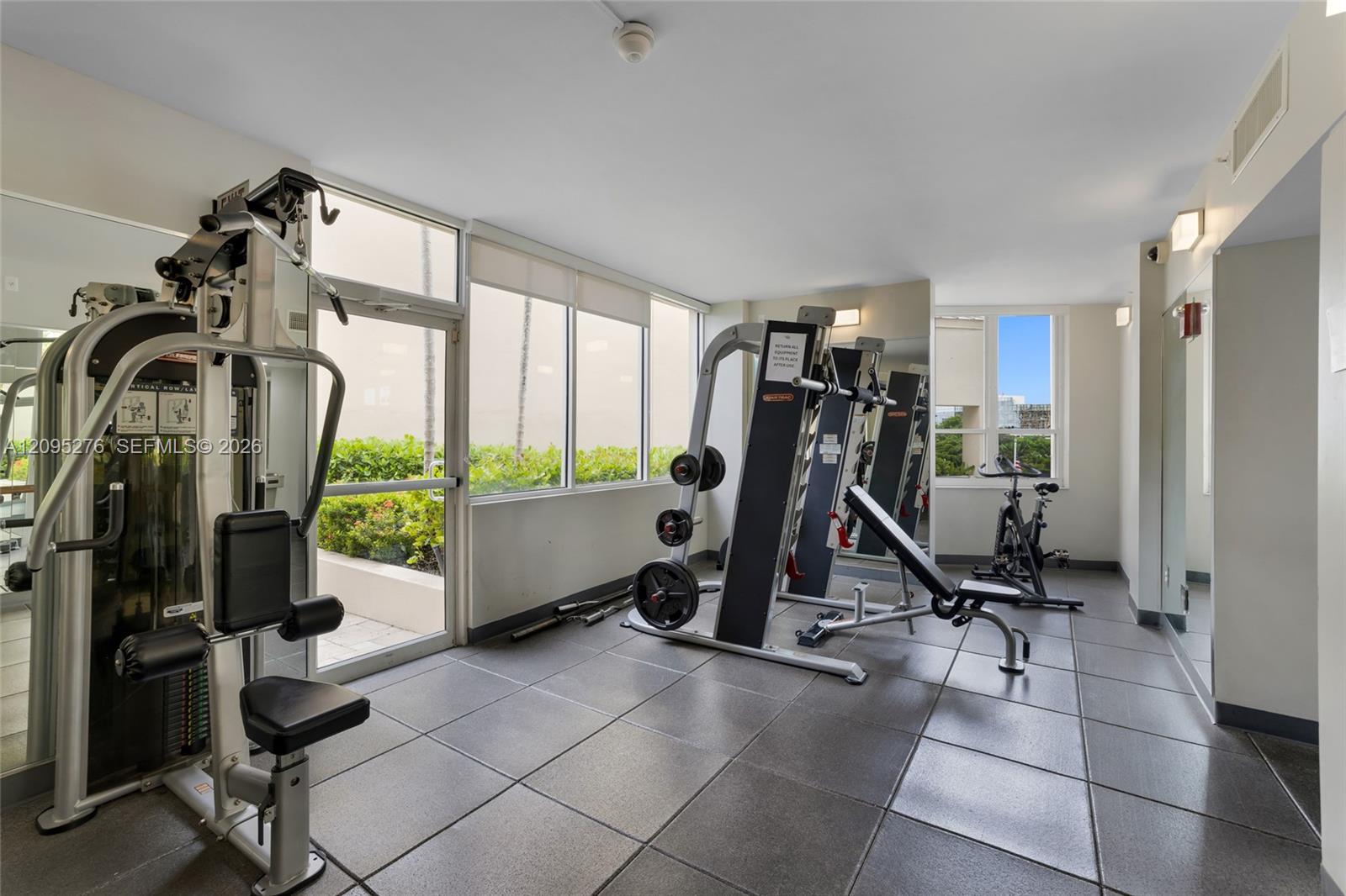 Condominium fitness room with weight machines, a stationary bike, and large windows overlooking greenery.