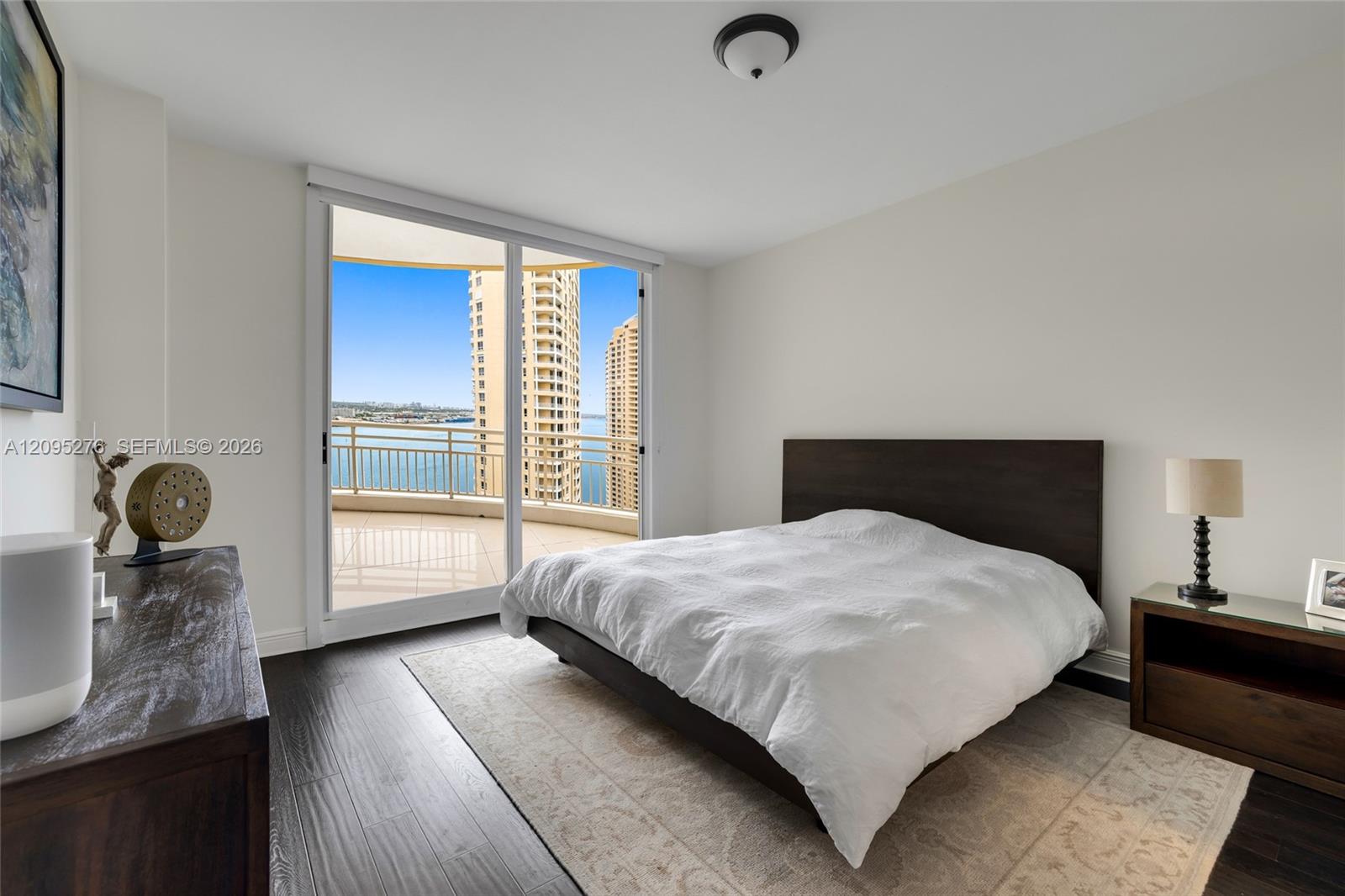 Bedroom with white bedding and sliding doors opening to a balcony overlooking the water and high-rises.