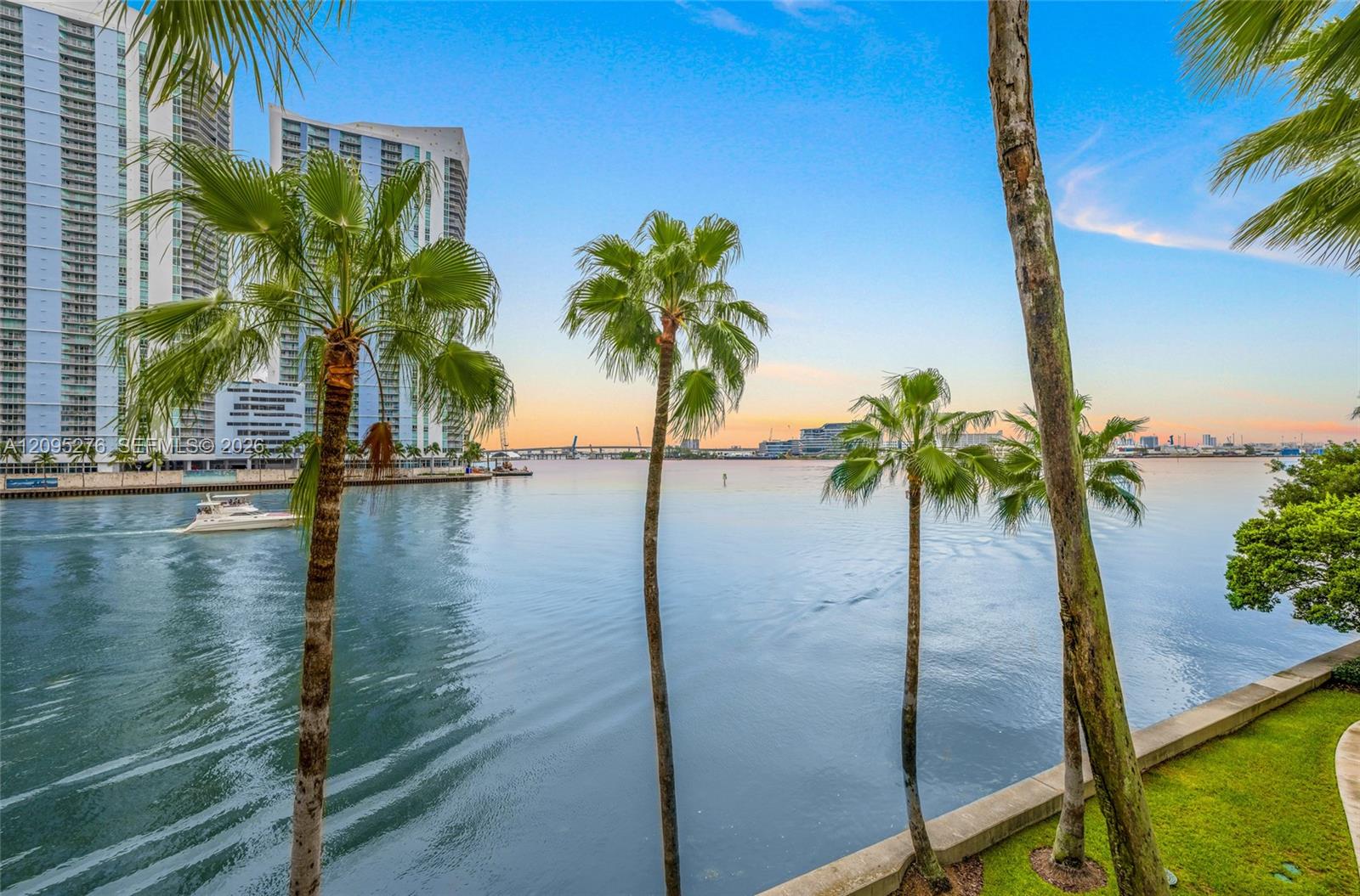 Waterfront view framed by palm trees, with high-rises, a passing boat, and the city skyline across the bay.